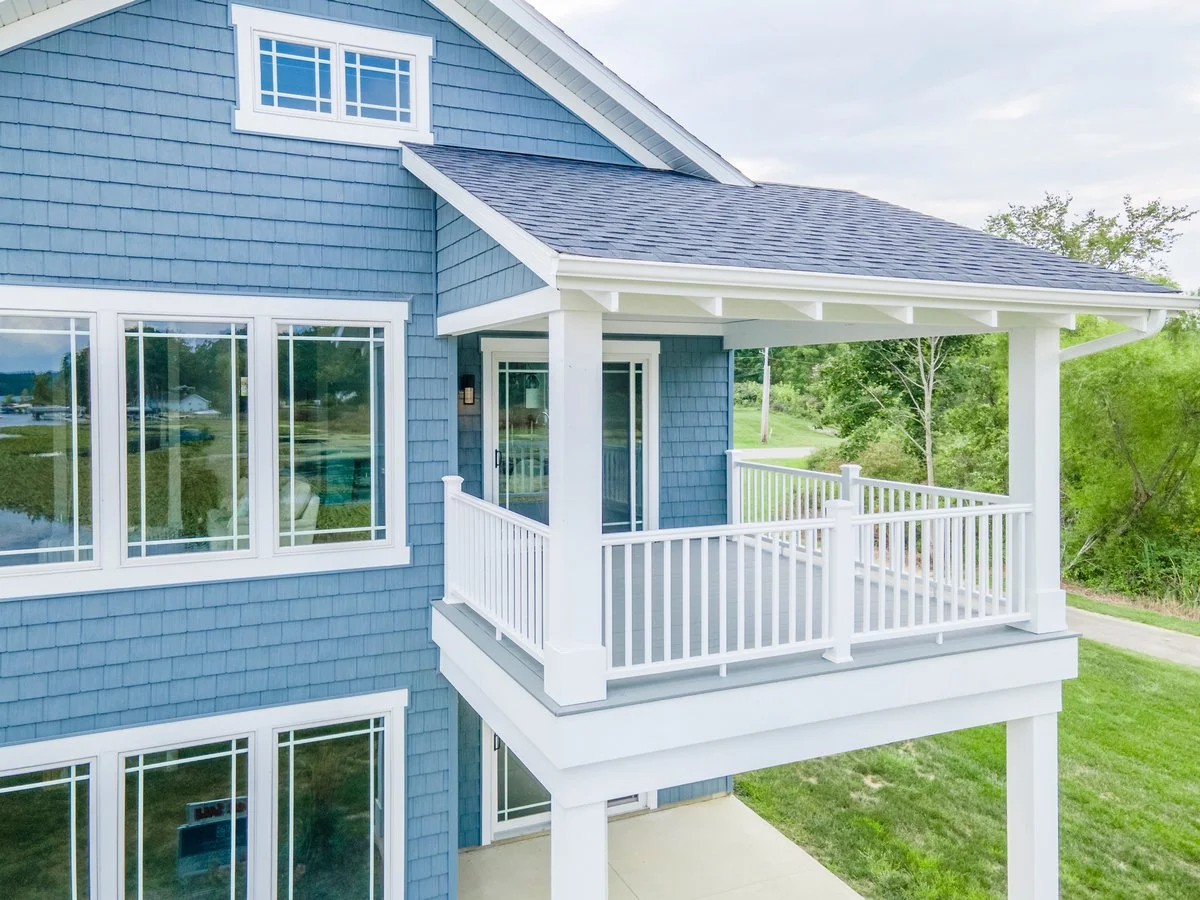 A blue house with a small covered balcony, white railing, and large windows, surrounded by green trees and grass.