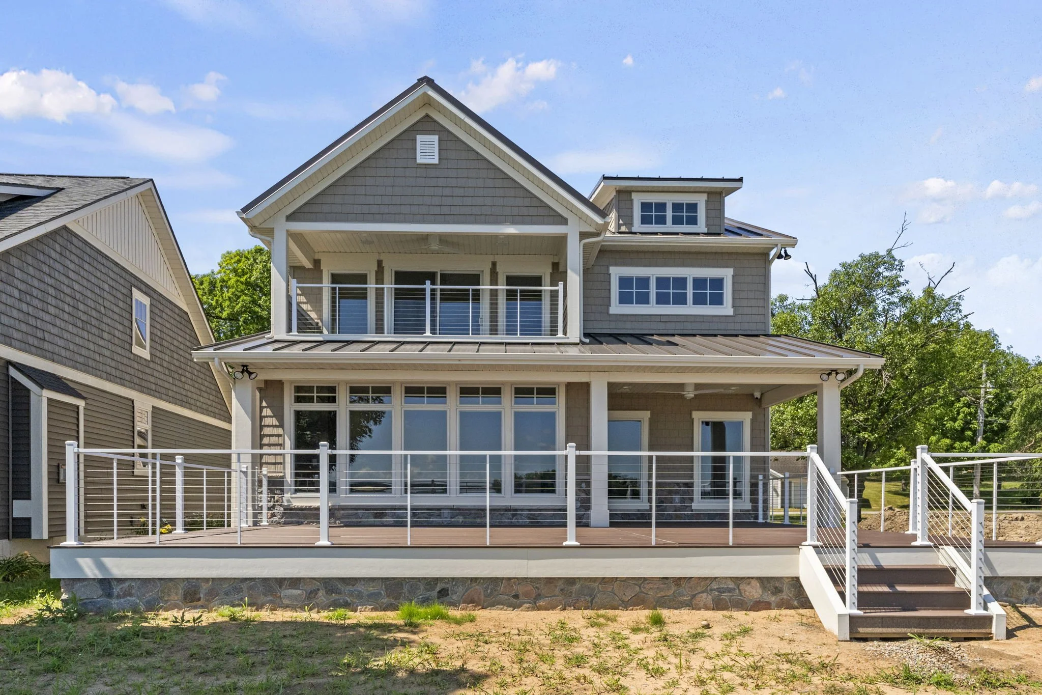 New two-story house with gray siding, large front windows, and a wooden deck with white railings, built on a stone foundation, with a staircase leading down to the yard, under a partly cloudy blue sky.