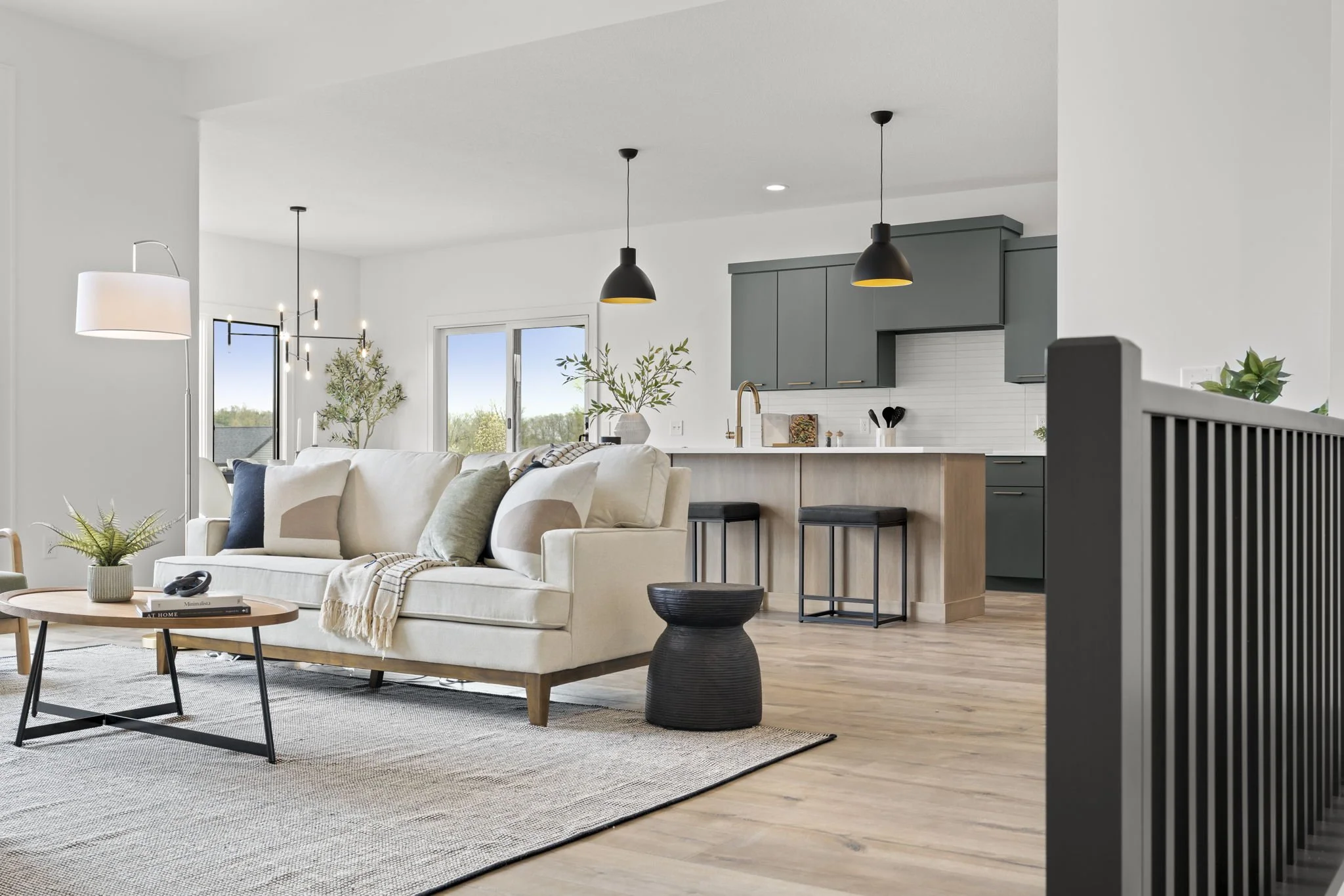 Modern open-concept living room and kitchen with white walls, wooden flooring, a beige sofa with pillows, a round coffee table, black pendant lights, an island with black stools, gray cabinets, and minimal decor.