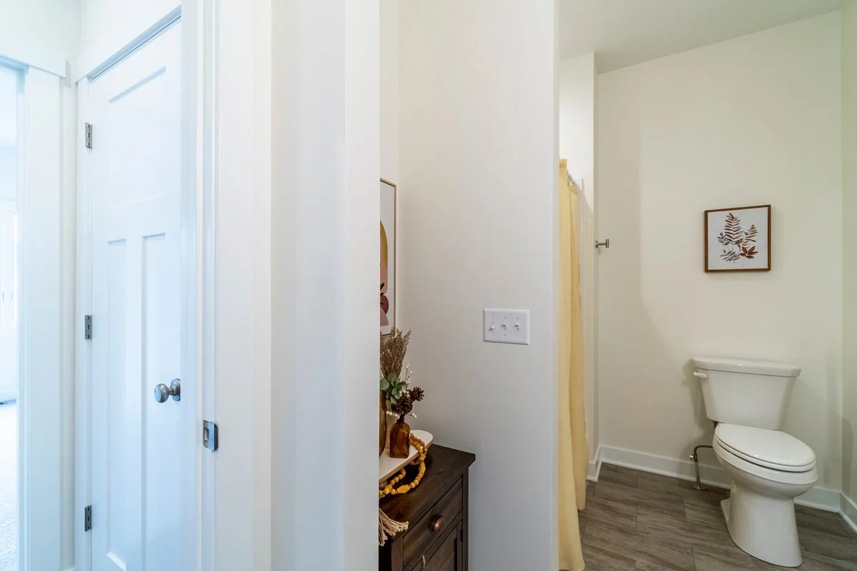 Bathroom with a toilet, framed artwork of leaves, light-colored walls, and wood-style flooring.