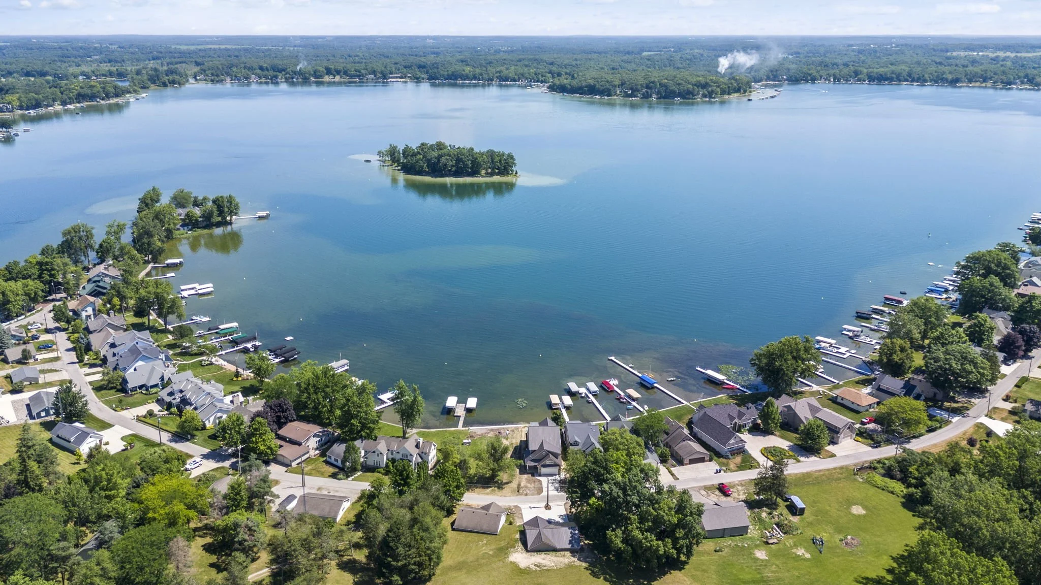 Aerial view of a lake with houses and docks along the shoreline, surrounded by lush green trees and a small island in the water.