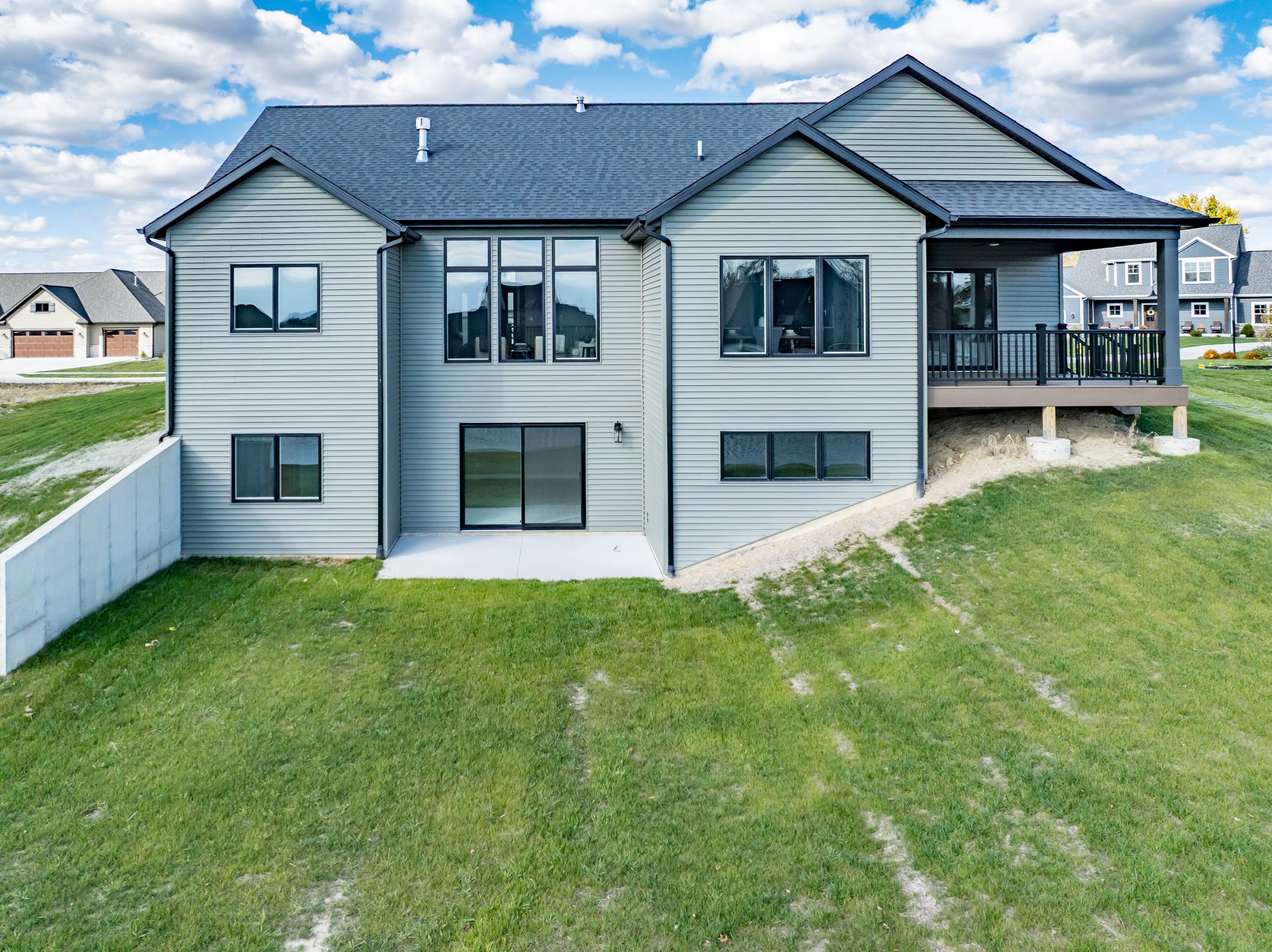 Back of a gray two-story house with a deck and large windows, set in a green yard with a concrete patio and neighboring houses in the background.