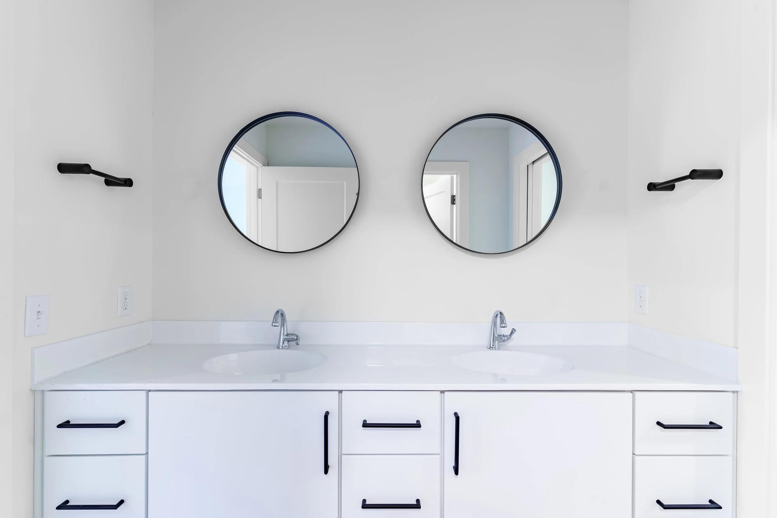 Bathroom vanity with two sinks, two oval mirrors, black fixtures, and a white cabinet.