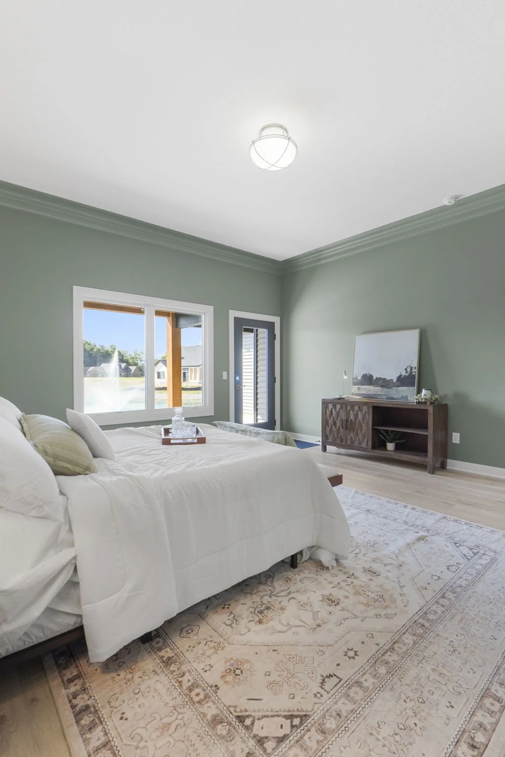 A bedroom with a white bed, cream pillows, a beige patterned rug, a light green wall, a window, a black door, and a wooden console with a mirror on top.