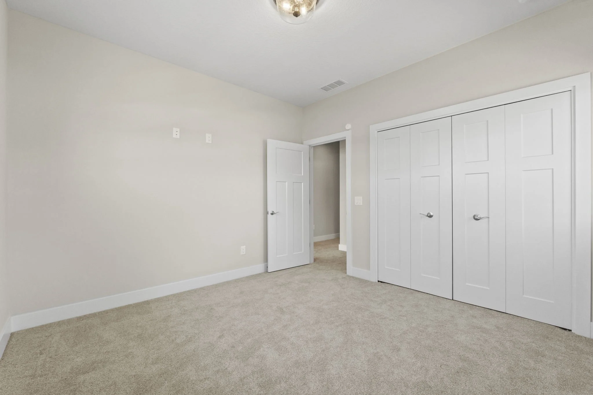 Empty bedroom with beige carpet, white walls, a closed closet with double doors, and an open door leading to a hallway.