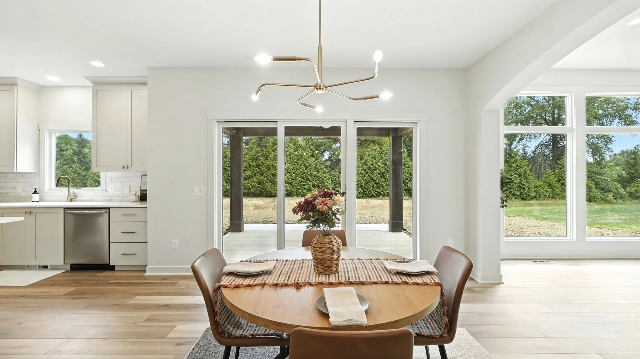 Dining area with a round wooden table, four brown chairs, a flower vase, and a table runner, situated in a bright open-concept space with large windows and sliding glass doors showing an outdoor patio and green trees.