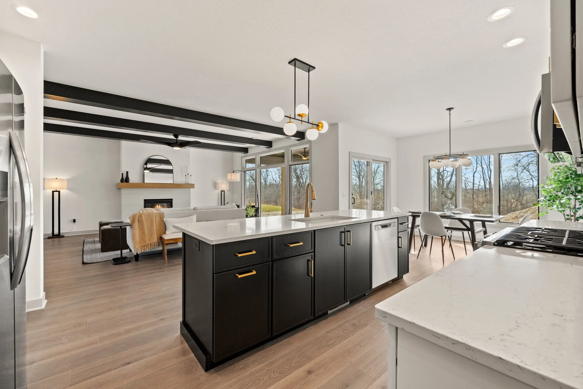 Open-concept kitchen and living area with black kitchen island, white countertops, modern lighting fixtures, large windows, and a view of trees outside.