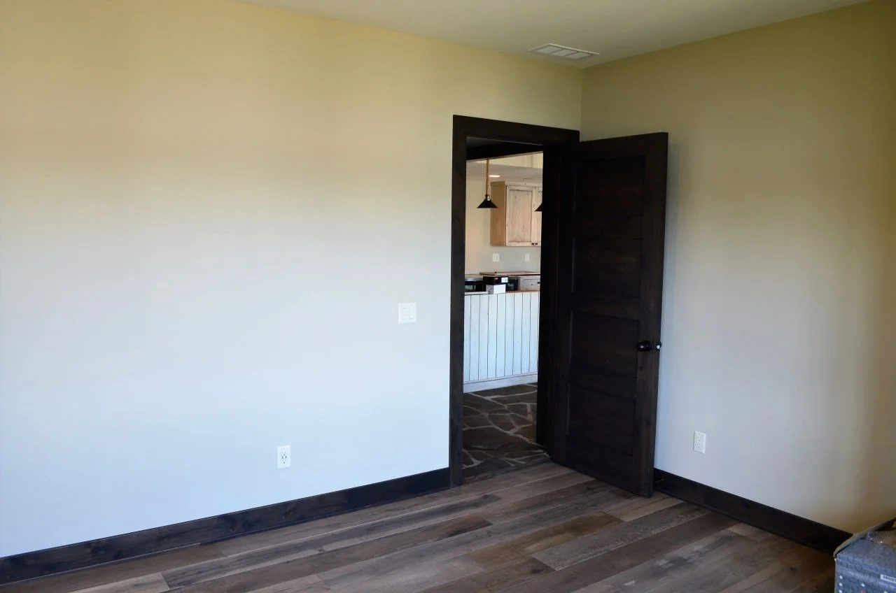 Empty room with white walls, dark wooden baseboards, a partially open dark wooden door, and a view into a kitchen with wooden cabinets and pendant lights