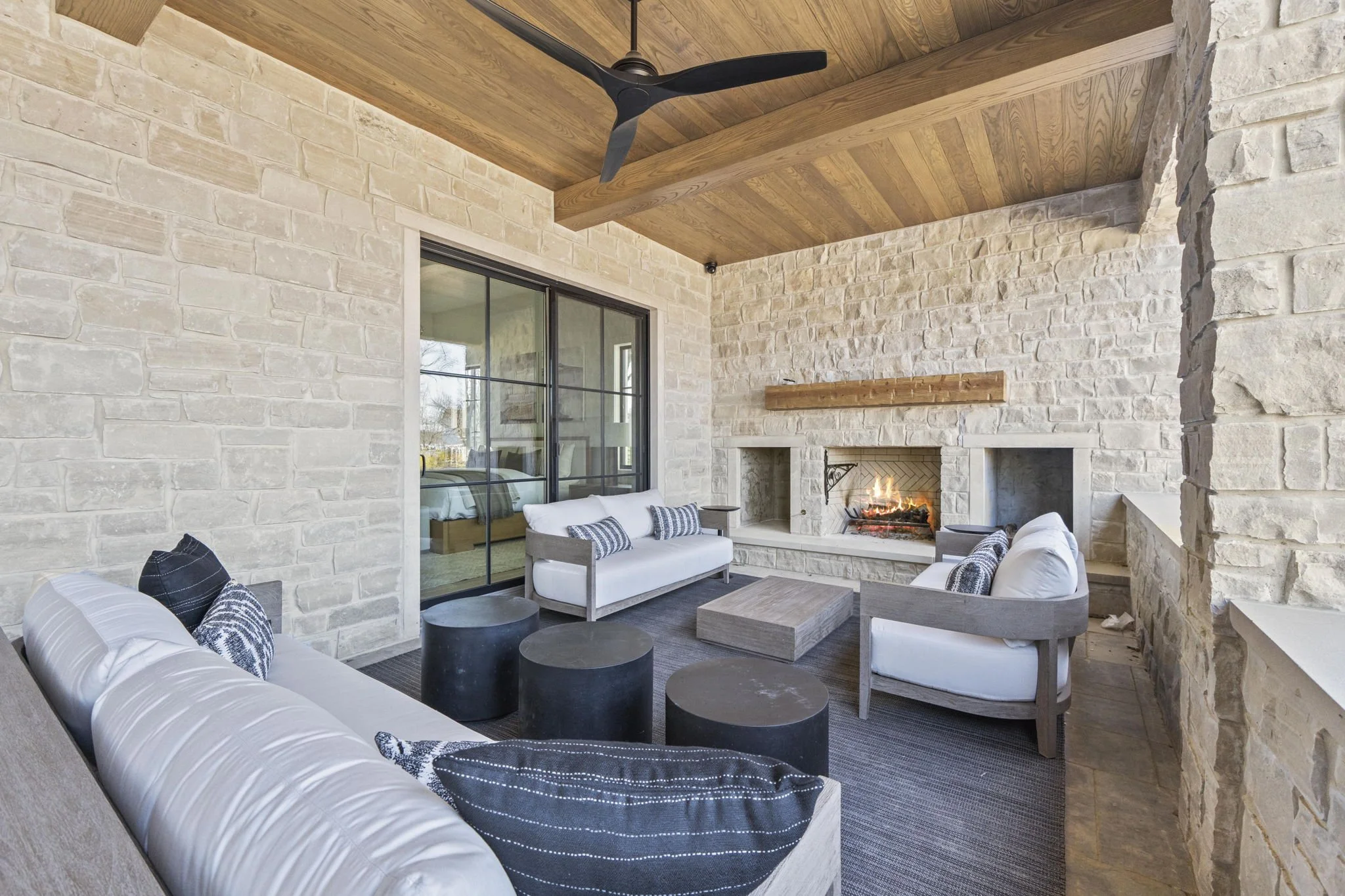 A cozy living room with cream-colored brick walls, a fireplace with a fire burning, a wooden beam on the ceiling, black ceiling fan, and white and gray furniture, including sofas and chairs, arranged around a coffee table.