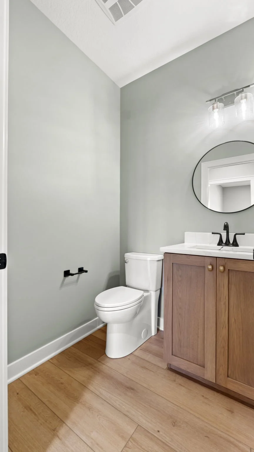 A modern bathroom with a toilet, wooden vanity with a white countertop, black faucet, round mirror, light fixture, and light gray walls.