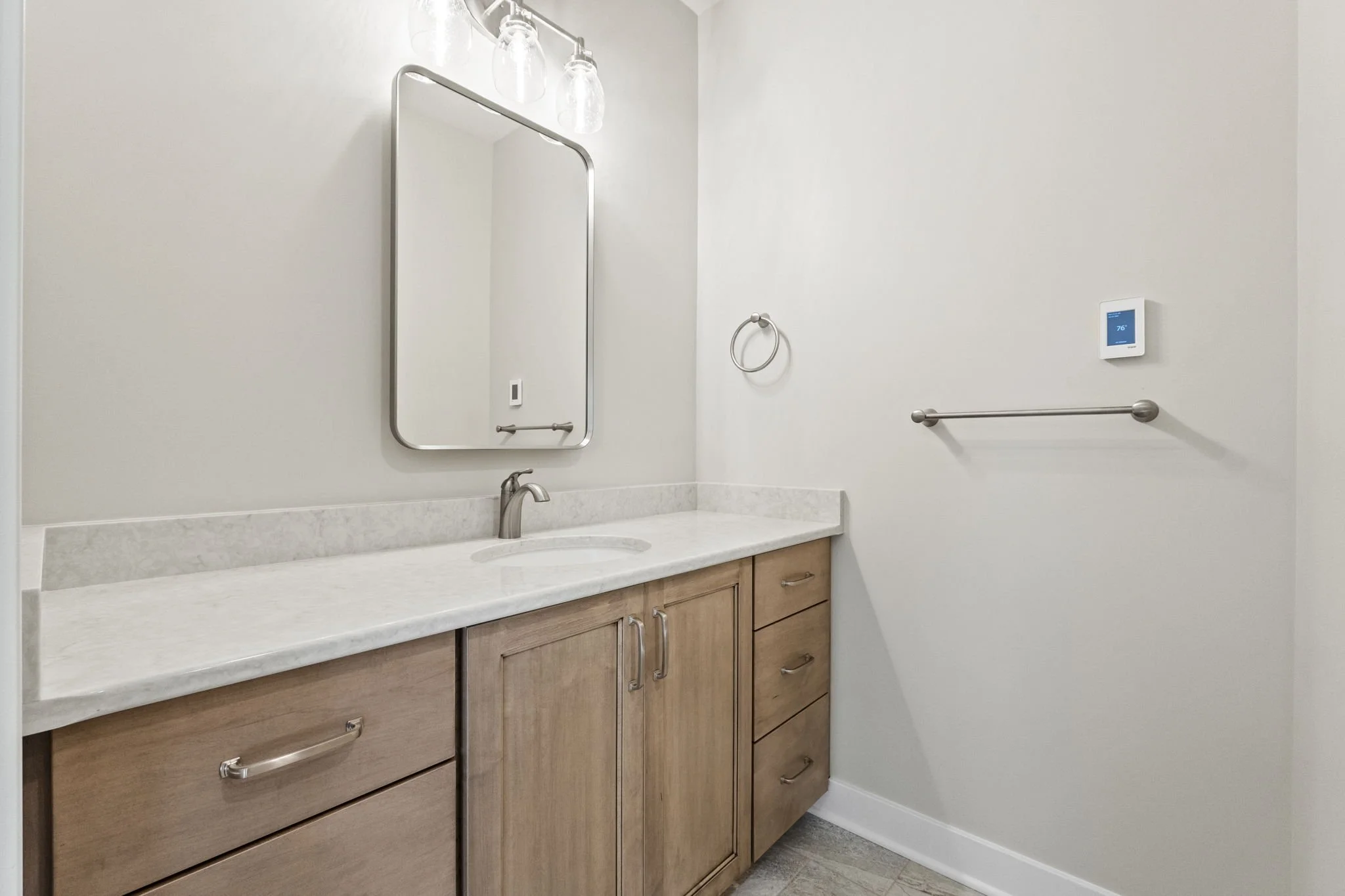 Empty bathroom vanity with wooden cabinets, marble countertop, faucet, large mirror, towel ring, towel bar, and thermostat on the wall.