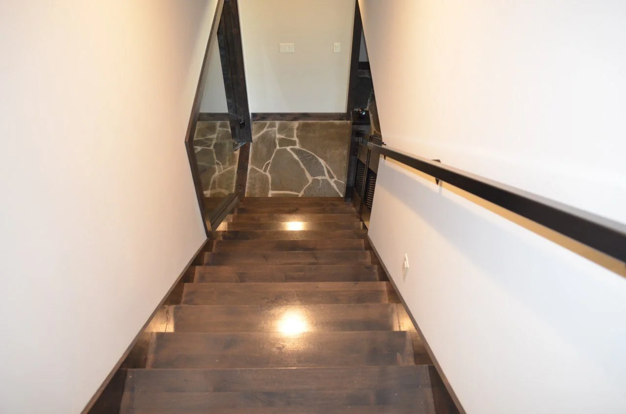 A view looking down a staircase with dark wooden steps, black metal handrails on both sides, white walls, and a stone floor at the bottom.