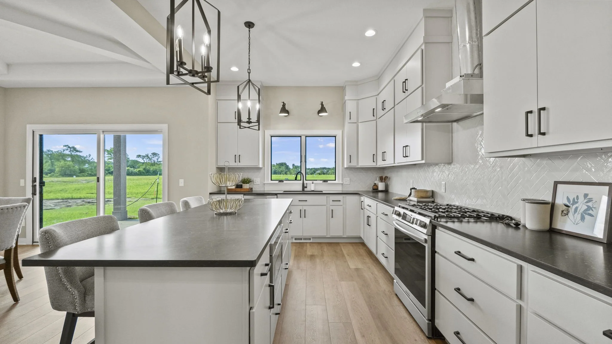 Modern kitchen with white cabinets, black handles, wood flooring, and a large island counter. Light fixtures hang above the island, and there are large windows showing a green outdoor landscape.