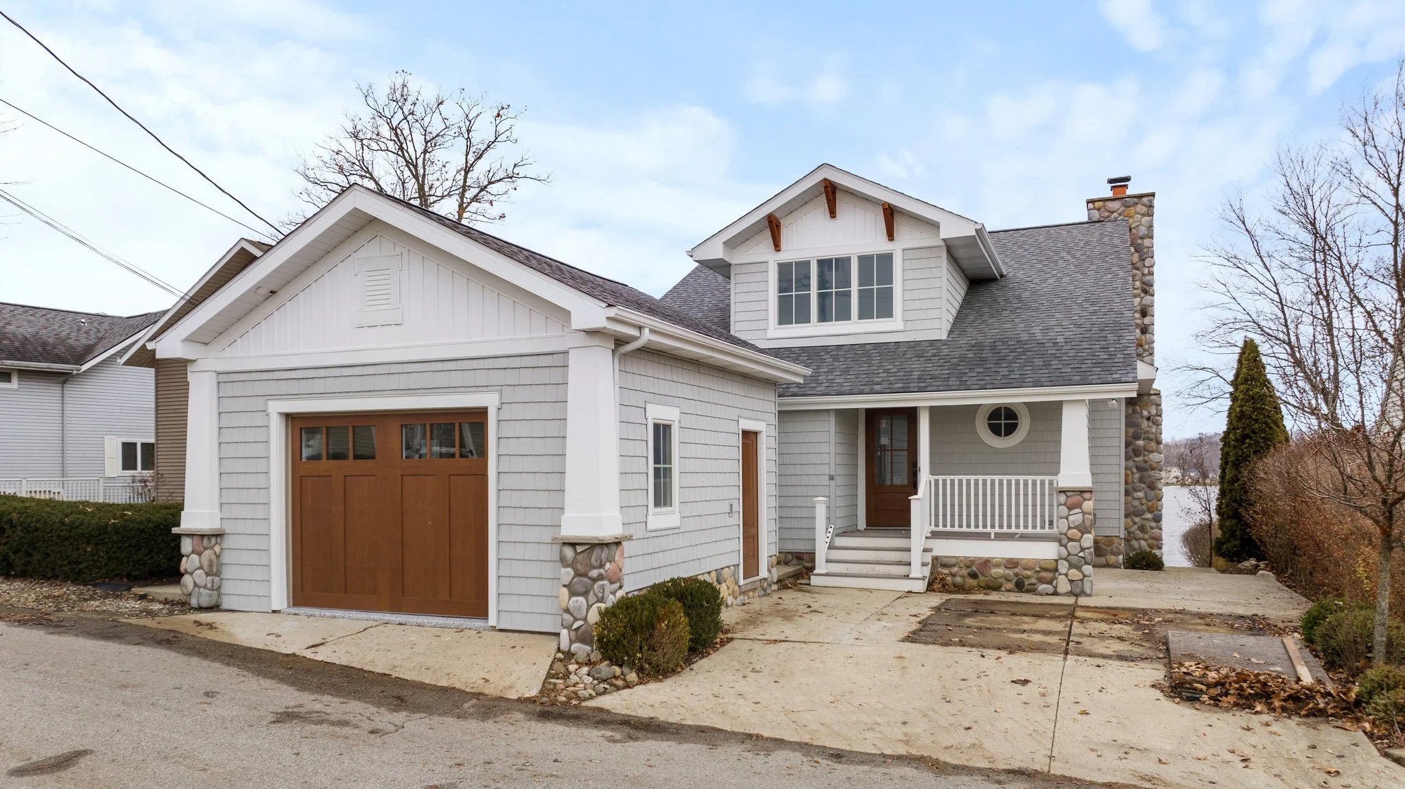 Front view of a house with gray siding, stone accents, brown garage door, and small front porch with white railing, set on a partly cloudy day.