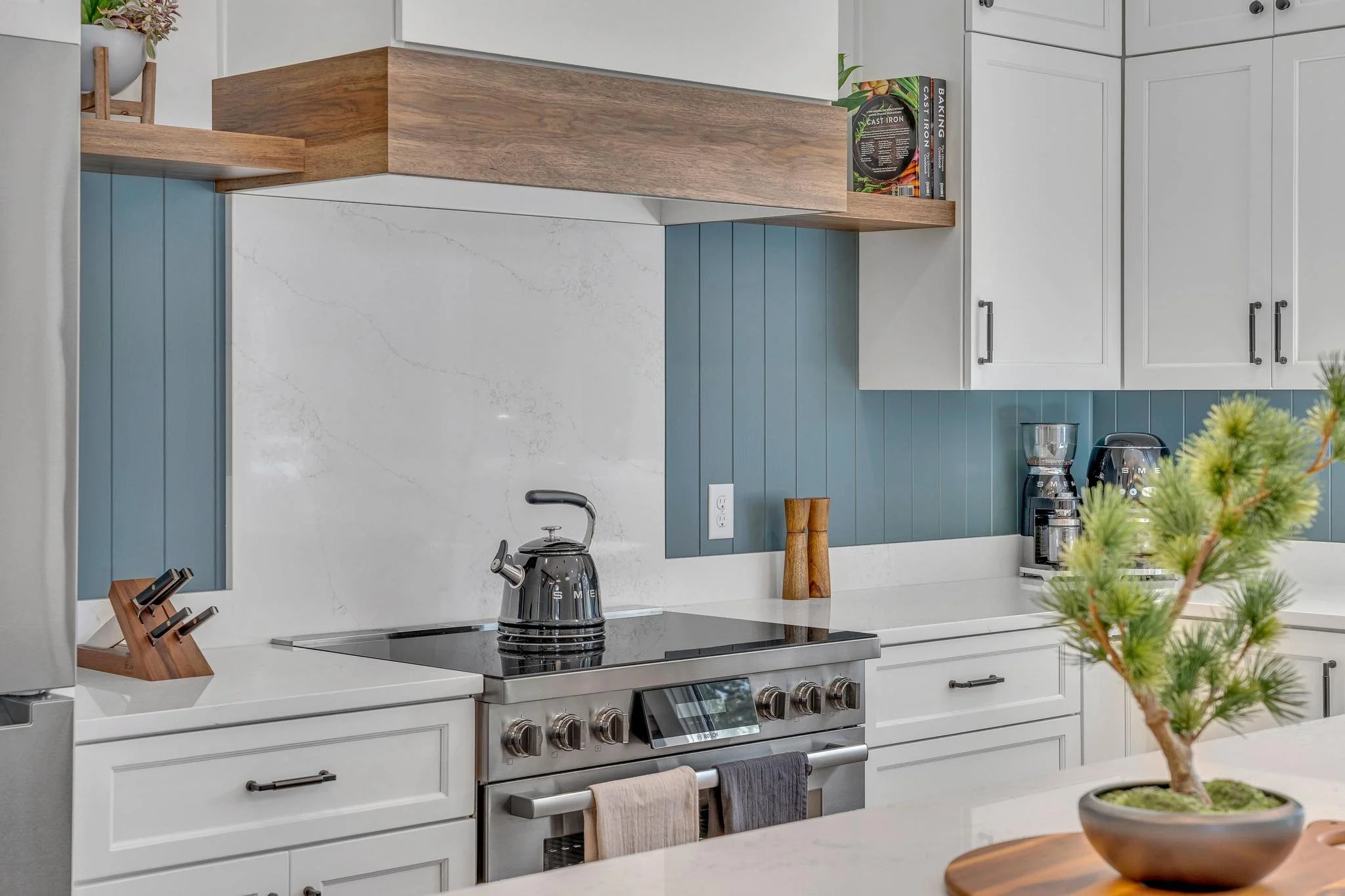 Modern kitchen with white cabinets, a white marble backsplash, and blue wooden paneling. There are black appliances, including a kettle on the stove. Open shelves hold books and small decor. A potted plant is on the counter in the foreground.