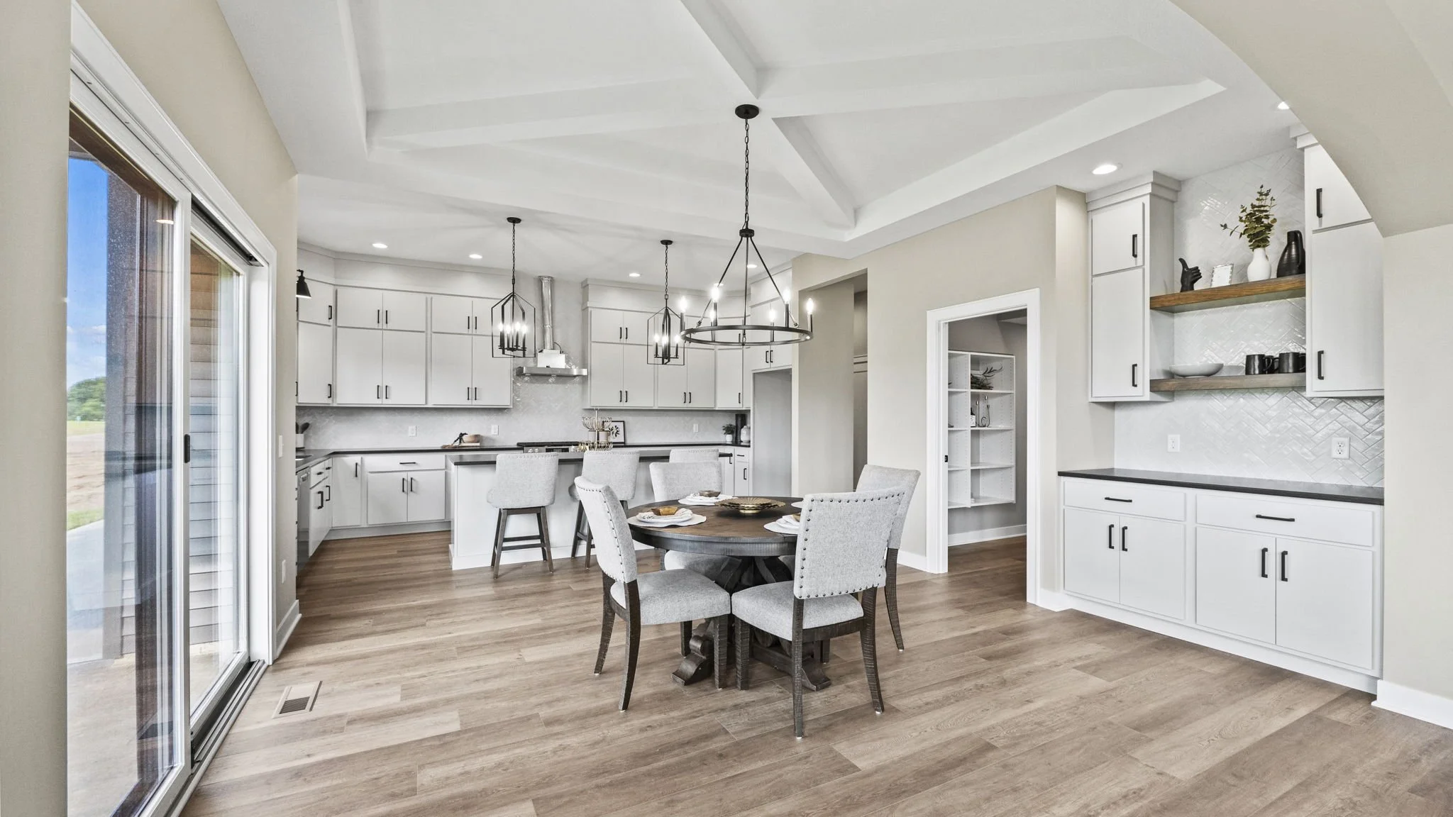 Open-concept kitchen and dining area with white cabinets, a round wooden table with upholstered chairs, hardwood floors, and modern black light fixtures.