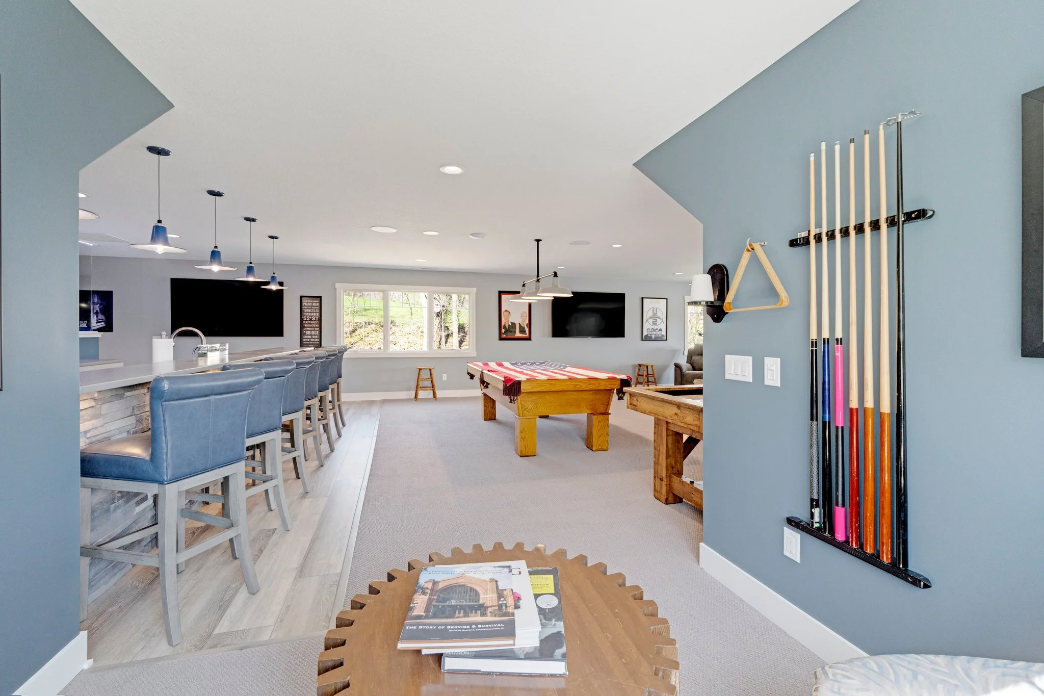 A spacious living area with a bar counter on the left with blue chairs, a pool table in the middle with an American flag cover, a wall-mounted TV, and a window showing trees outside. On the right, there are pool cues mounted on the blue wall and a sm