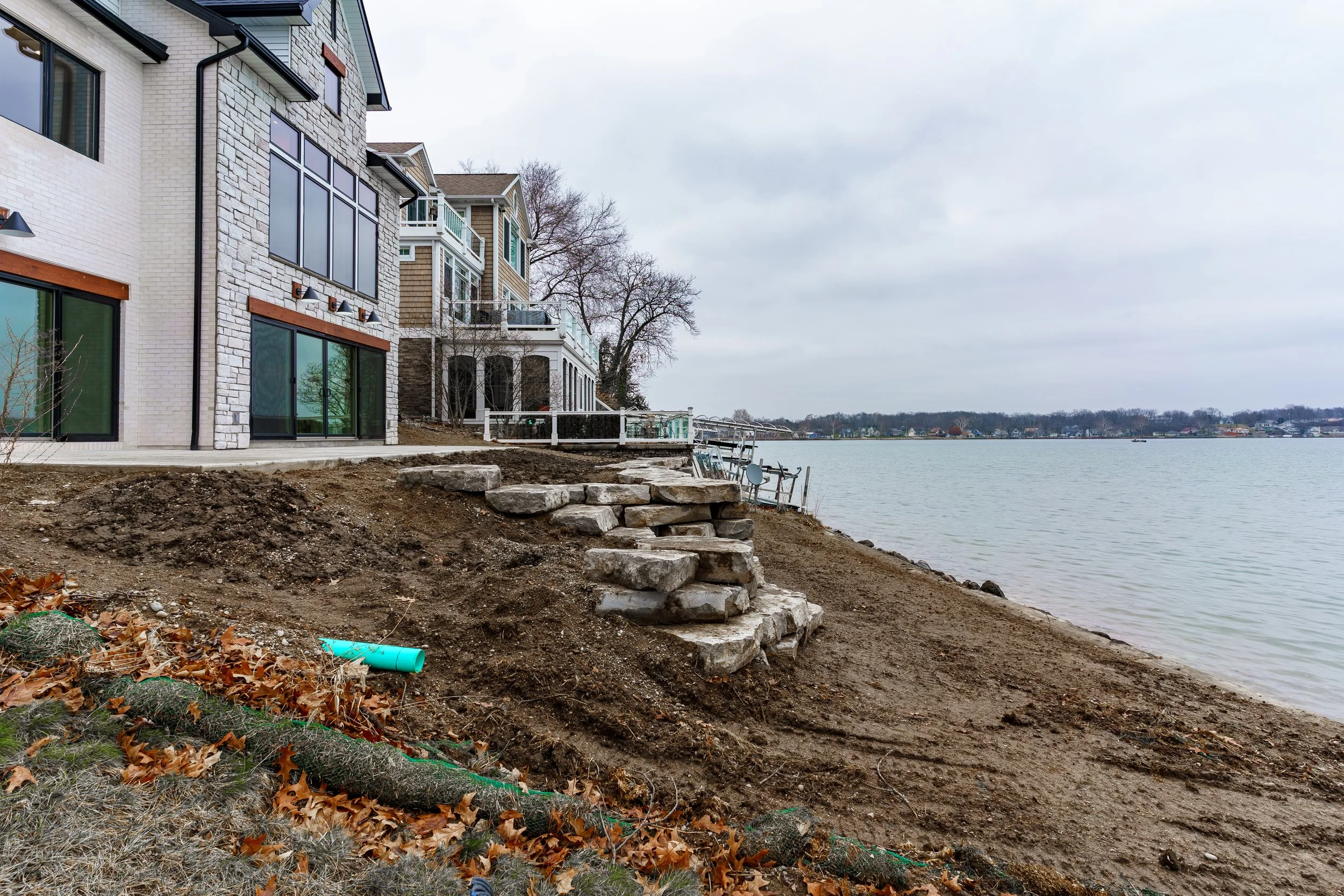 Multi-story residential building beside a body of water under an overcast sky, with a dirt shoreline and a set of stone steps leading to the water, construction debris in the foreground.