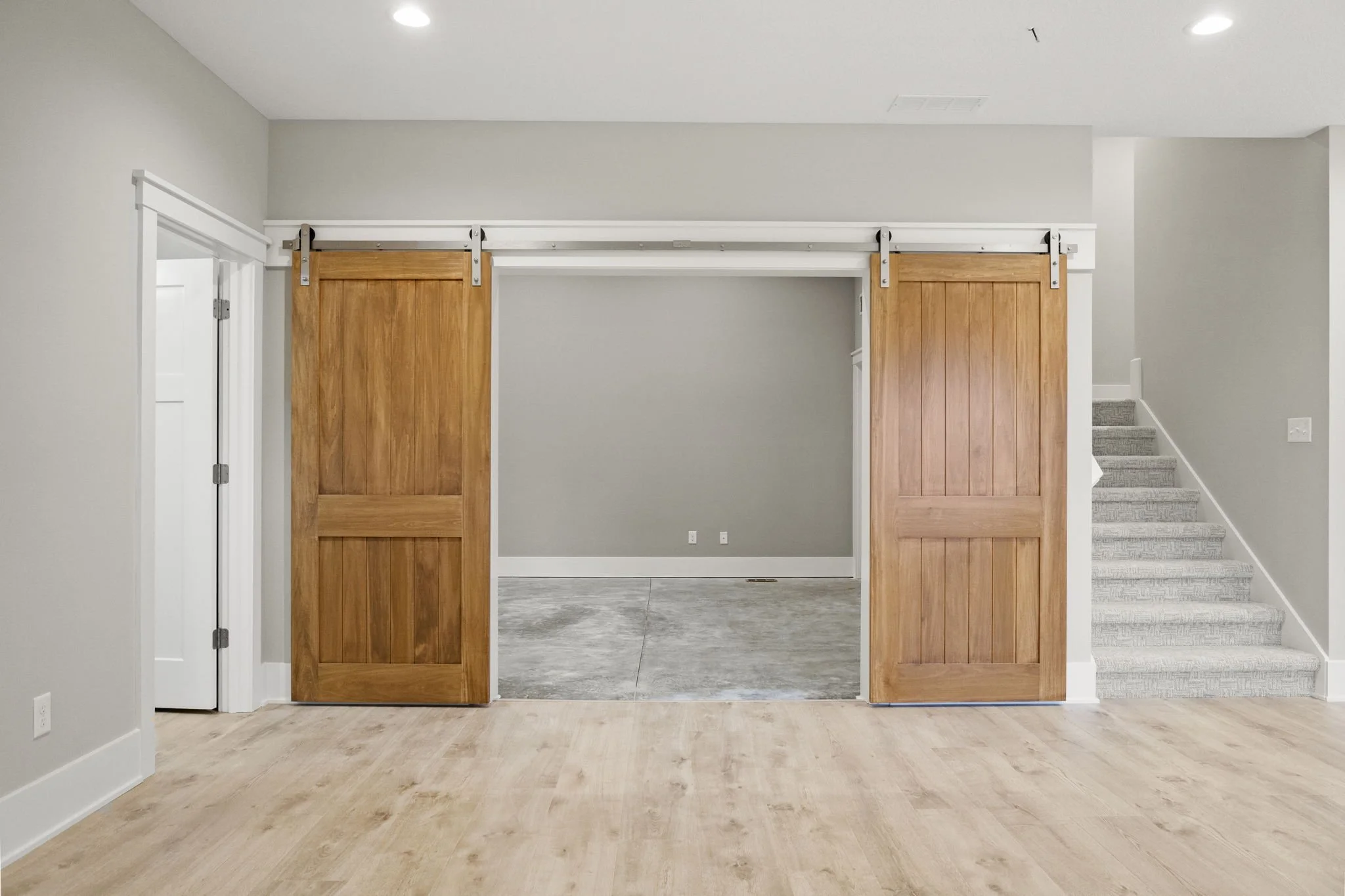Interior view of a room with light wood flooring, gray walls, and a sliding barn door made of wood. The barn door is partially open, revealing an adjacent room with concrete flooring. There are stairs with light carpeted steps on the right and a whit
