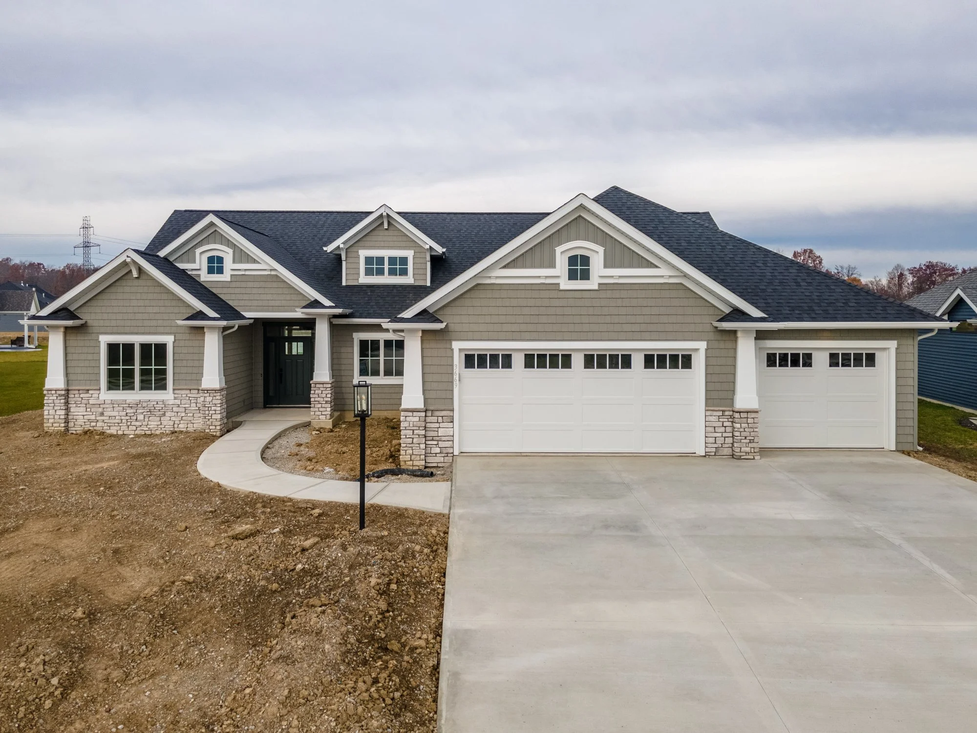 Newly constructed modern house with gray siding, stone accents, and a three-car garage, with a concrete driveway and a curved walkway leading to the front door, under a cloudy sky.