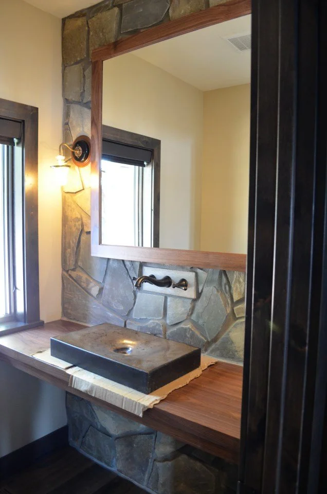 A rustic bathroom sink area with a unique stone and wood design, featuring a black vessel sink, a wall-mounted faucet, a mirror, and two windows with dark trim letting in natural light.