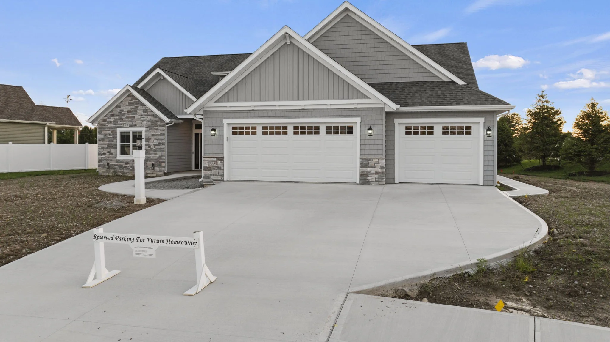 New house with gray siding, stone accents, three white garage doors, and a concrete driveway. A sign on the driveway reads "Reserved Parking For Future Homeowner." The house is surrounded by a partly landscaped yard with some grass and trees in the b