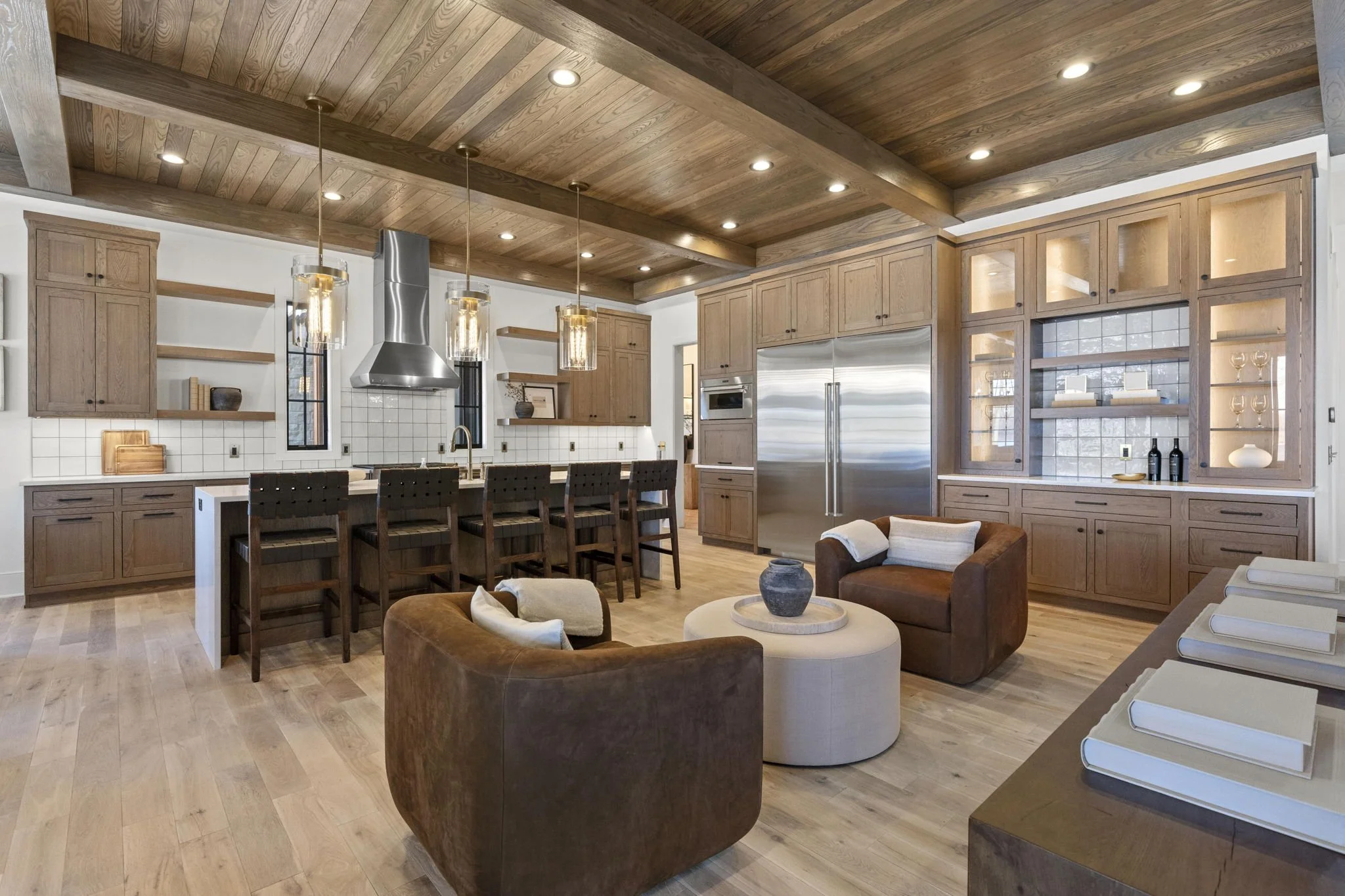 Open-concept kitchen and living room with wooden ceiling and cabinetry, stainless steel refrigerator, island with bar stools, and cozy seating area with brown chairs and a round ottoman.