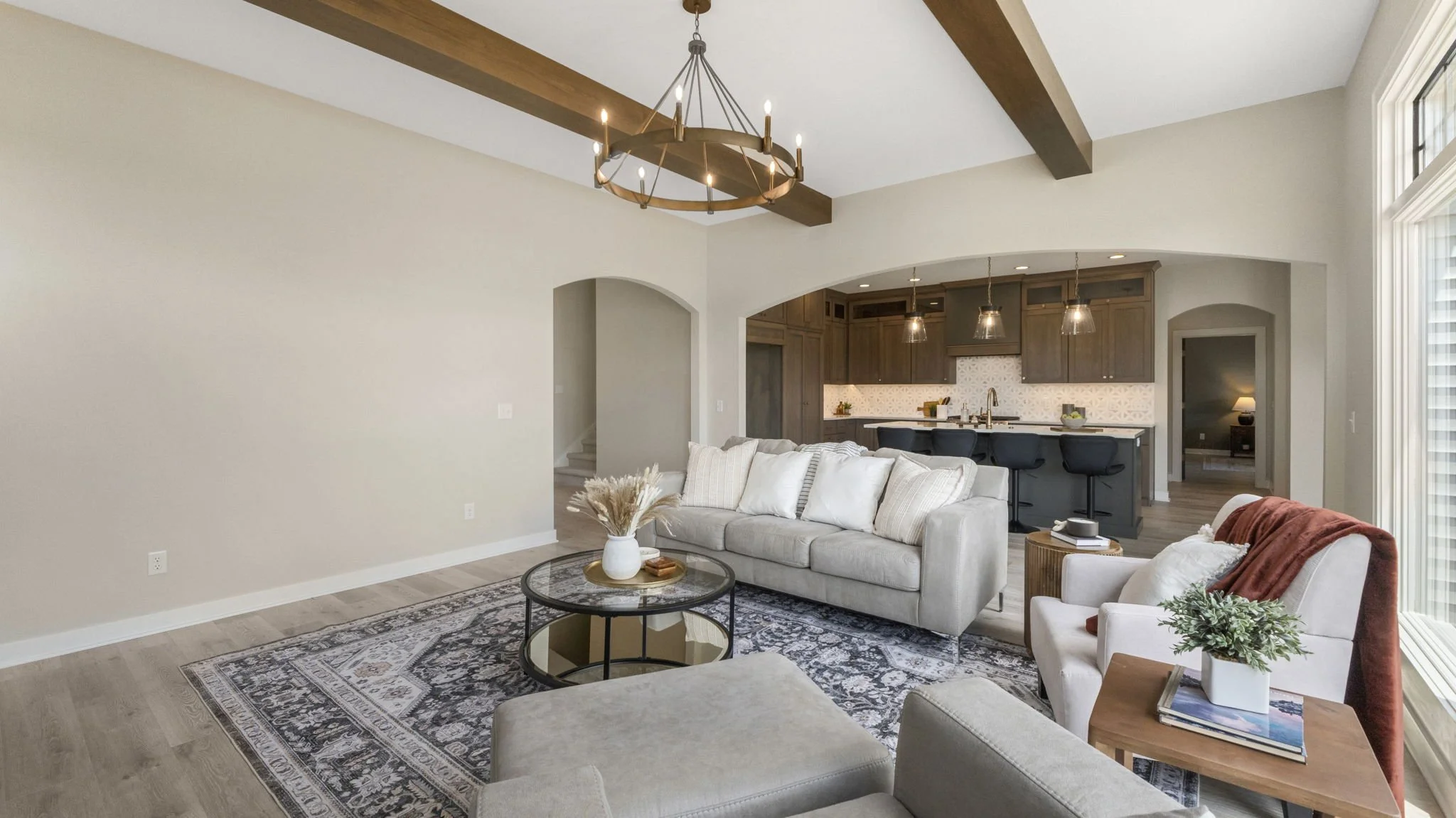Living room with a beige sofa, armchair with a red throw blanket, round coffee table with a white vase, and a patterned area rug. Open kitchen in the background with brown cabinets, pendant lights, and bar stools.