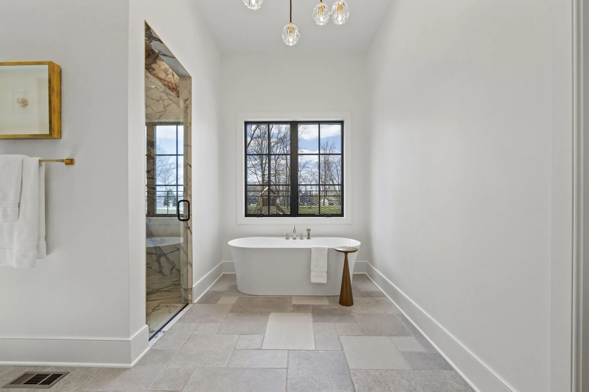 Modern bathroom with a freestanding bathtub beneath a window, adjacent to a walk-in shower with marble tile, white walls, and pendant lights.