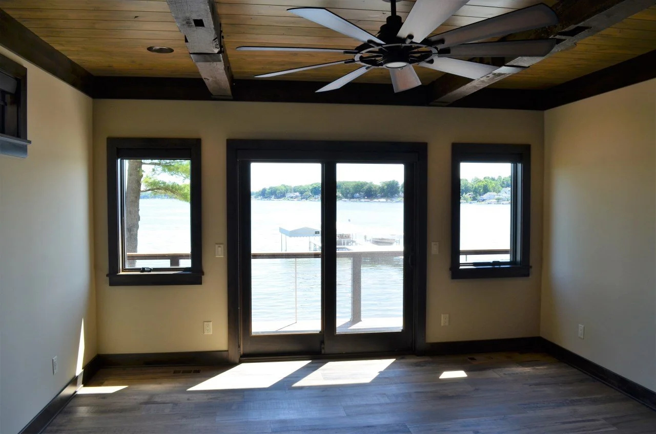 Empty room with a view of a lake or river through a glass sliding door and two windows, overlooking a dock and water, with a wooden ceiling and ceiling fan.