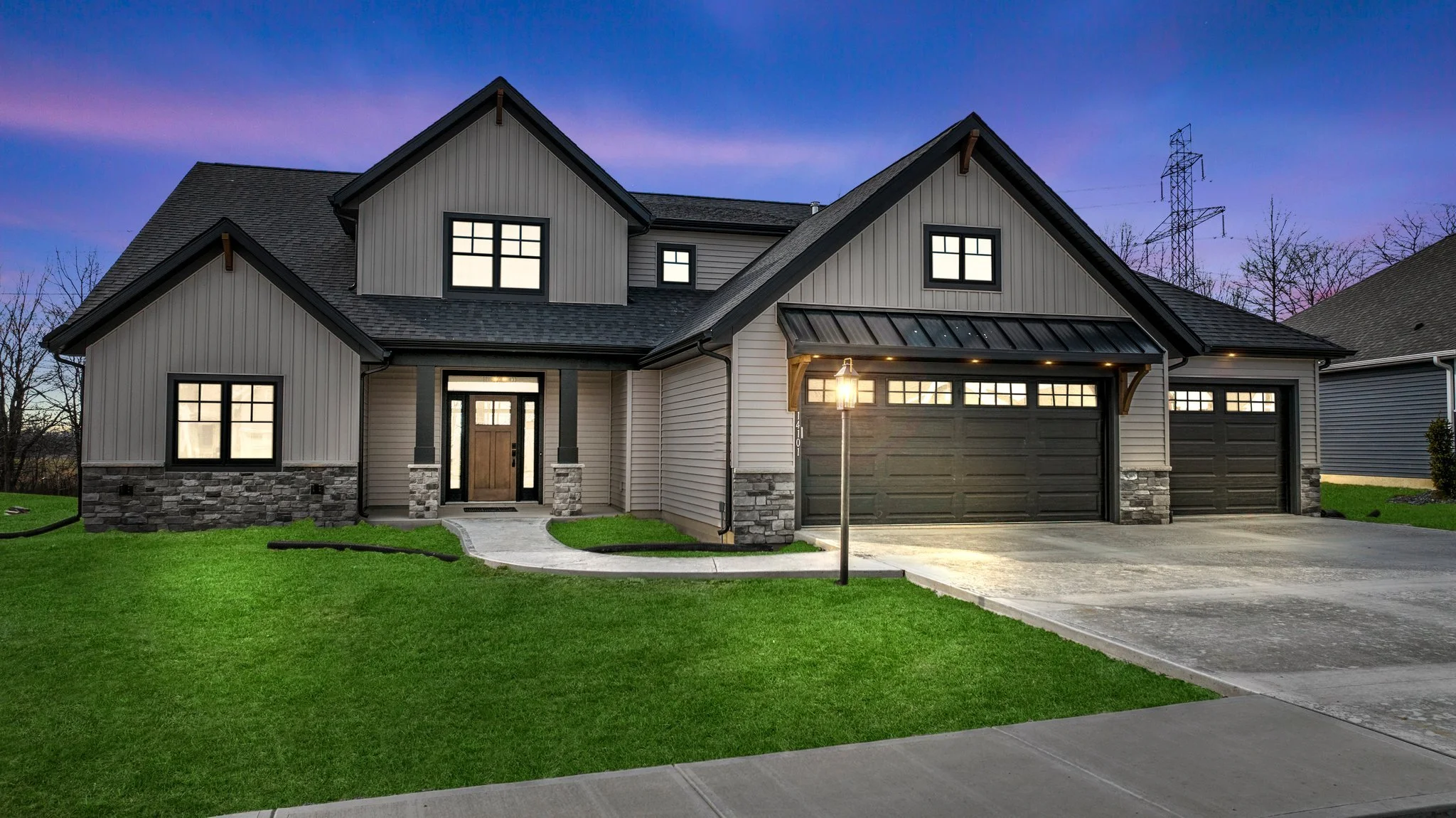 Modern two-story house with stone and beige siding, black trim, three garage doors, lit front porch, and a streetlamp outside during twilight.