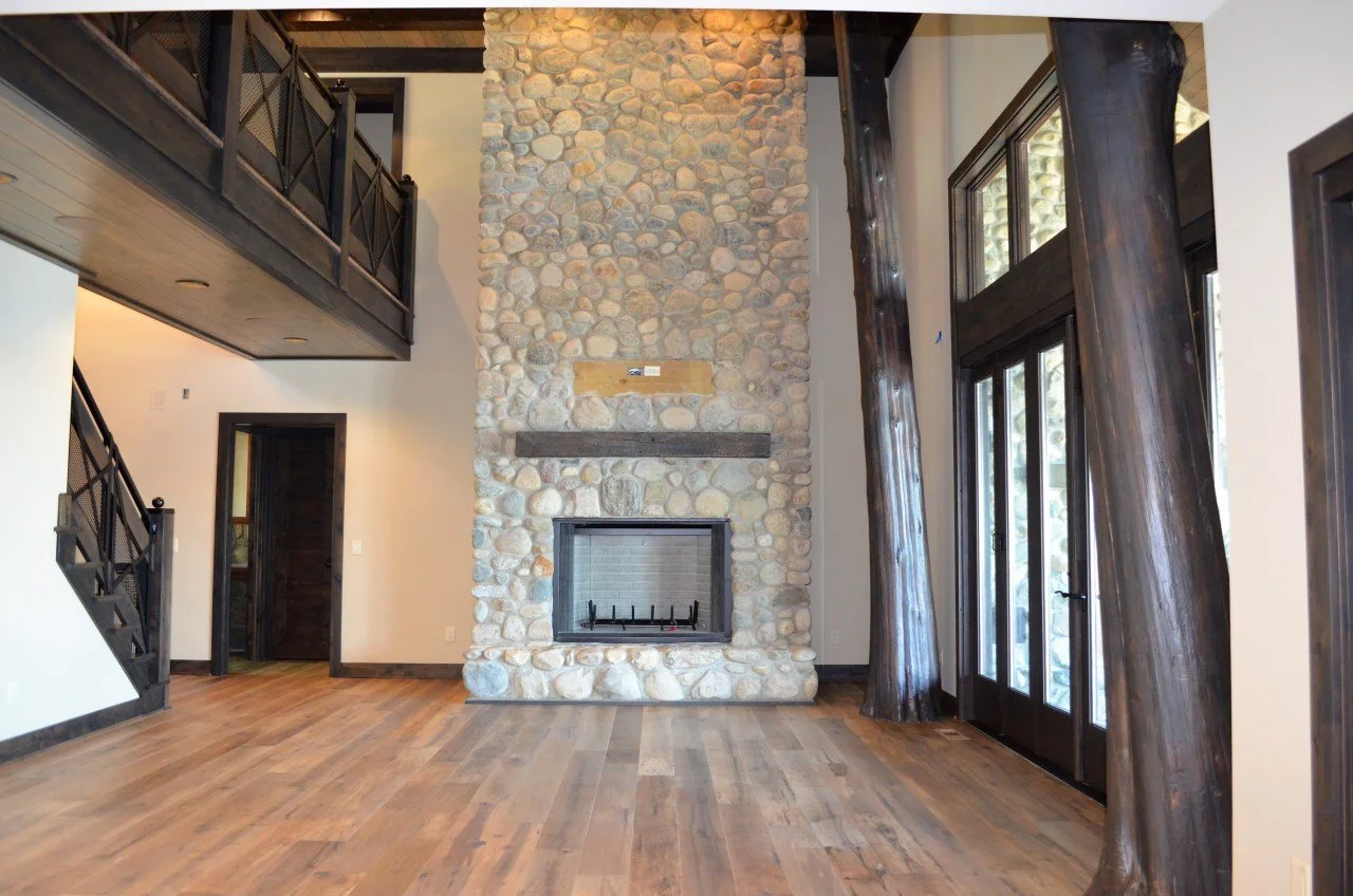 Living room with a large stone fireplace, wooden beams, wood flooring, and large glass windows and doors with tall wooden support beams.