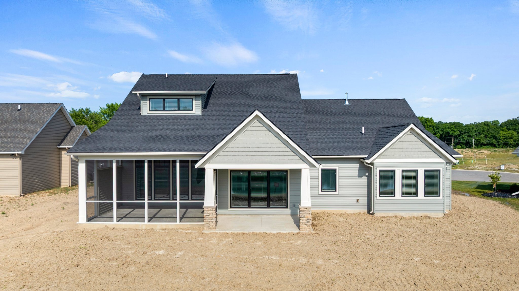 Back view of a modern house with a screened porch and multiple windows, set on a dirt lot with neighboring houses and green trees in the background.