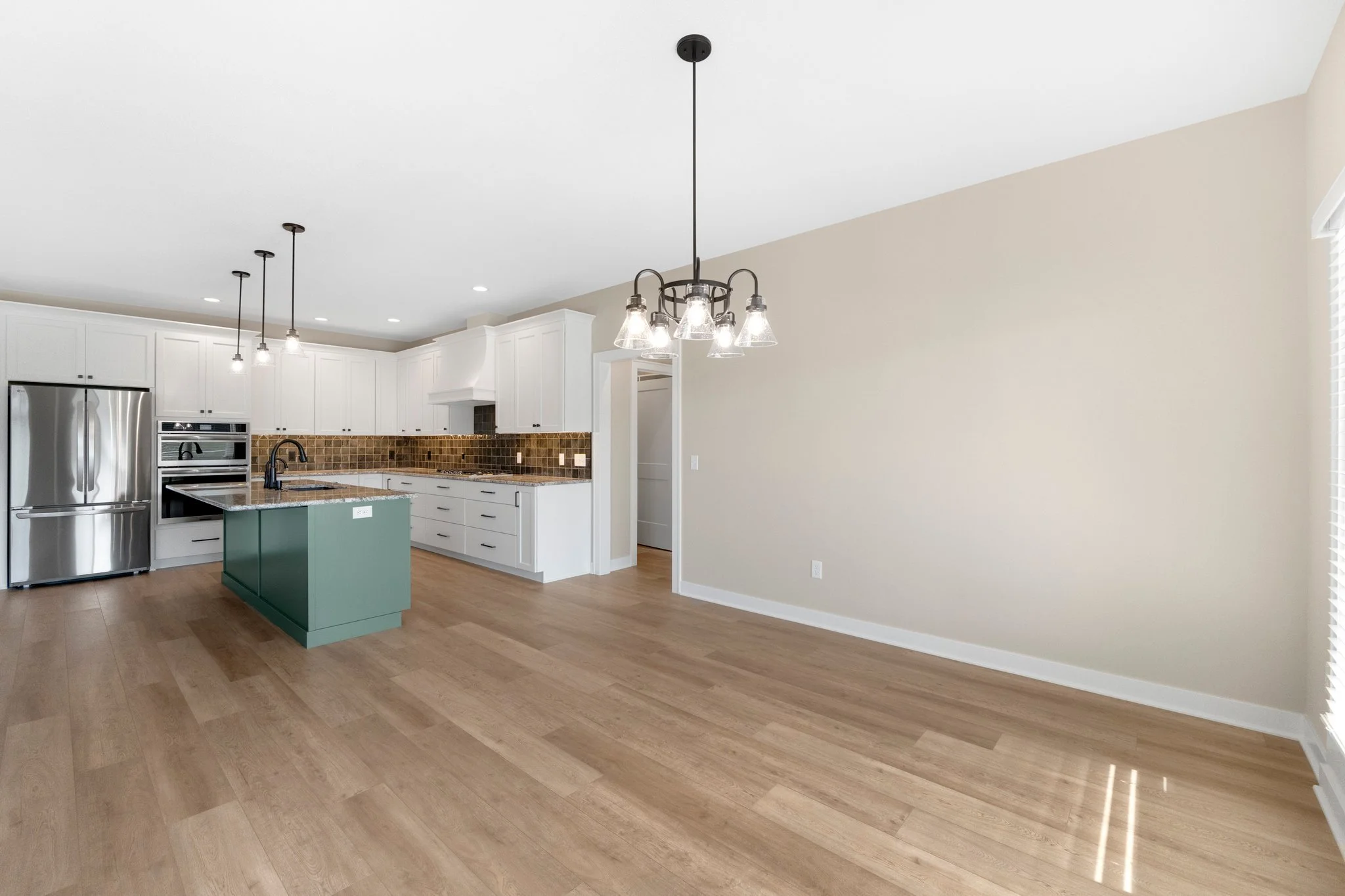 Empty modern kitchen and dining area with white cabinets, brown backsplash, wooden floor, and a green island in the center, no furniture.