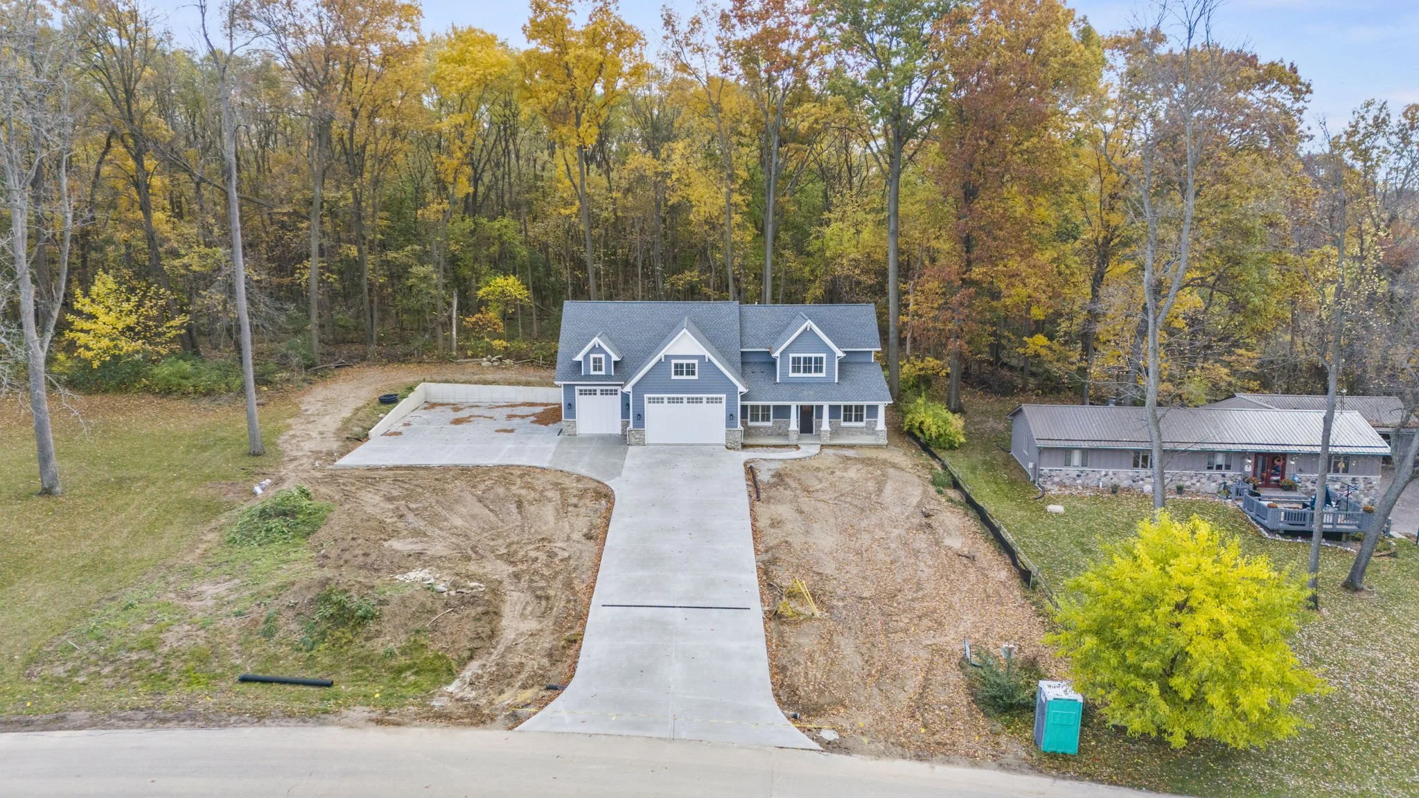 A new two-story house with a blue roof under construction, featuring a front driveway, in a suburban neighborhood with fall foliage.