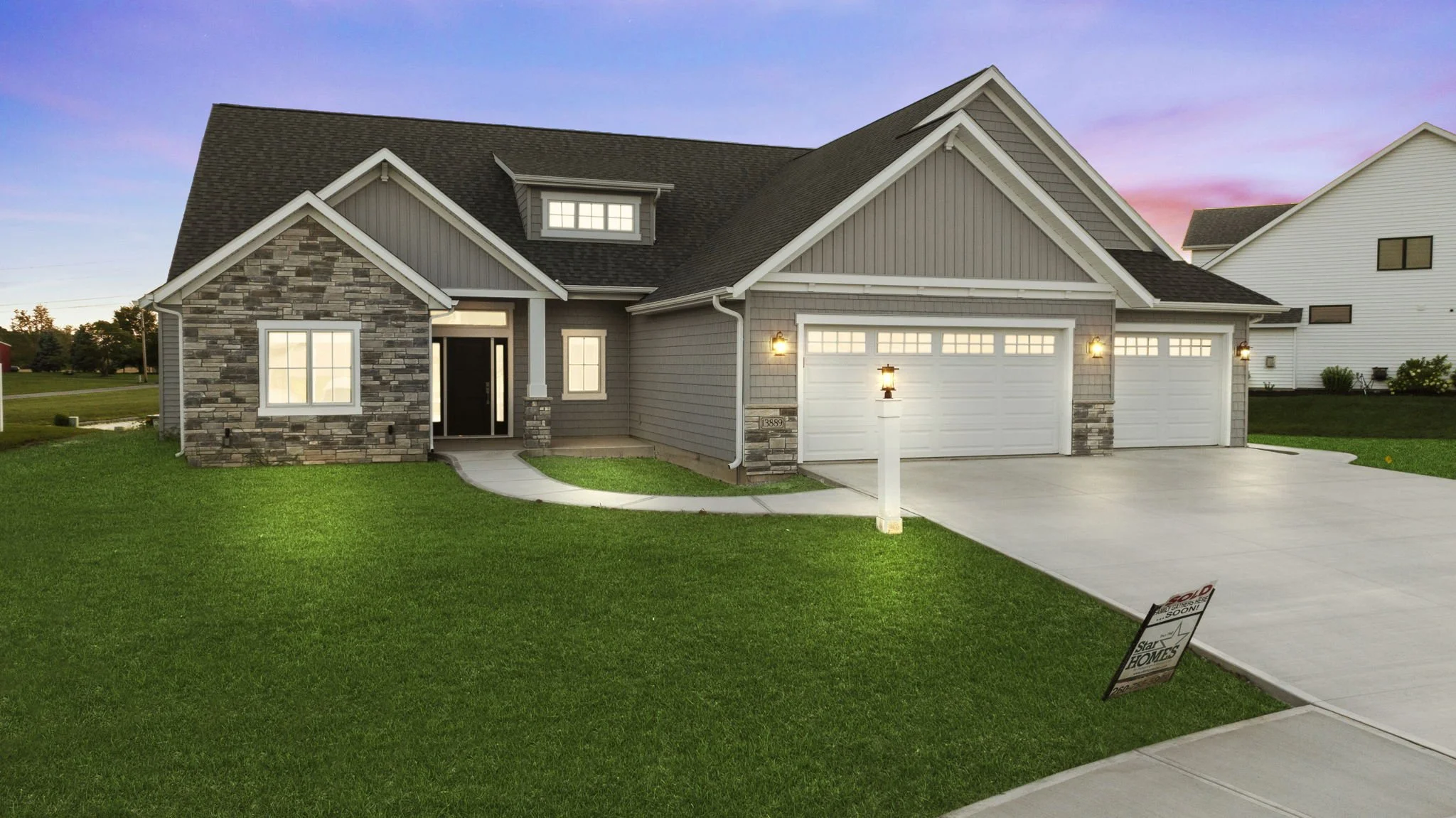 Newly built house with a gray exterior, stone accents, white garage doors, and a small front porch, illuminated by outdoor lights, with a well-manicured lawn and a sidewalk leading to the front door, during sunset.