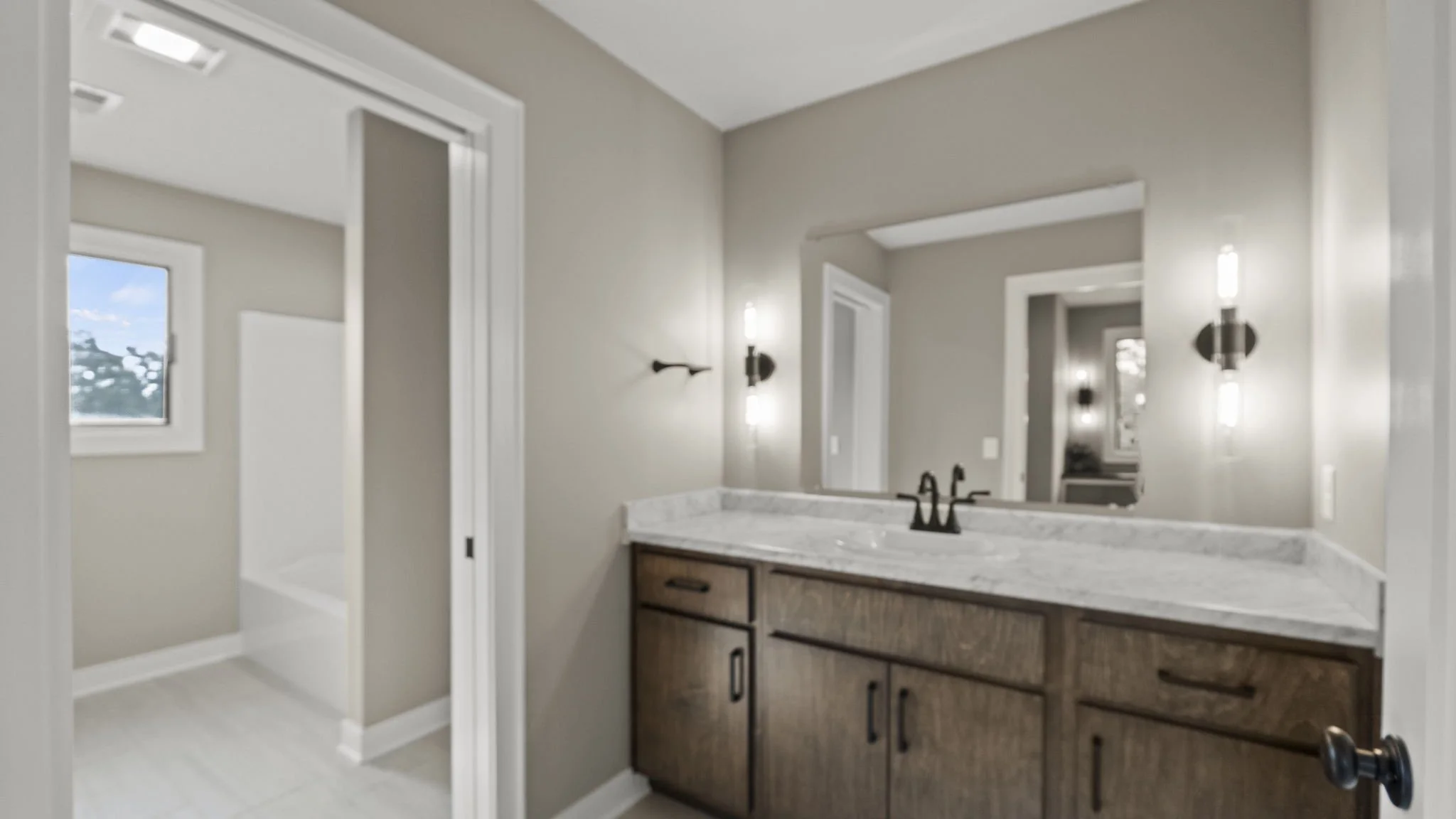 A bathroom with a wooden vanity, marble countertop, dual black faucets, large mirror, and modern black wall sconces on beige walls. There are two windows and a doorway leading to another room.