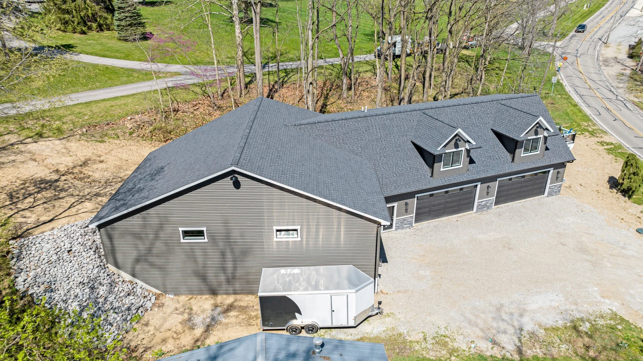 Aerial view of a residential house with a dark grey roof, attached garage, and a gravel driveway. The house is surrounded by trees and a patch of bare earth. There is also a small trailer parked near the house.