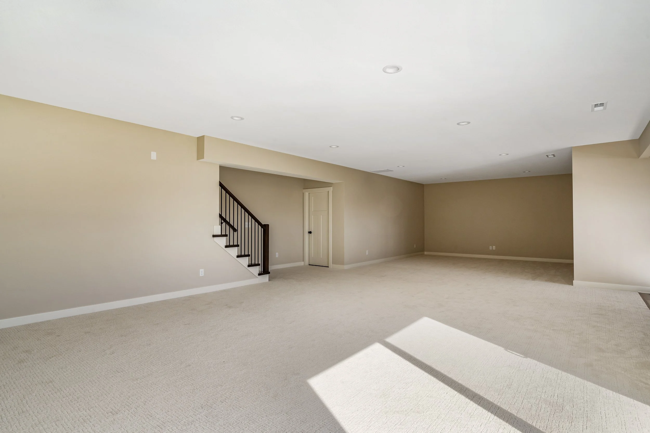 Empty spacious living room with beige walls, off-white carpet, and a staircase with dark wood railing.