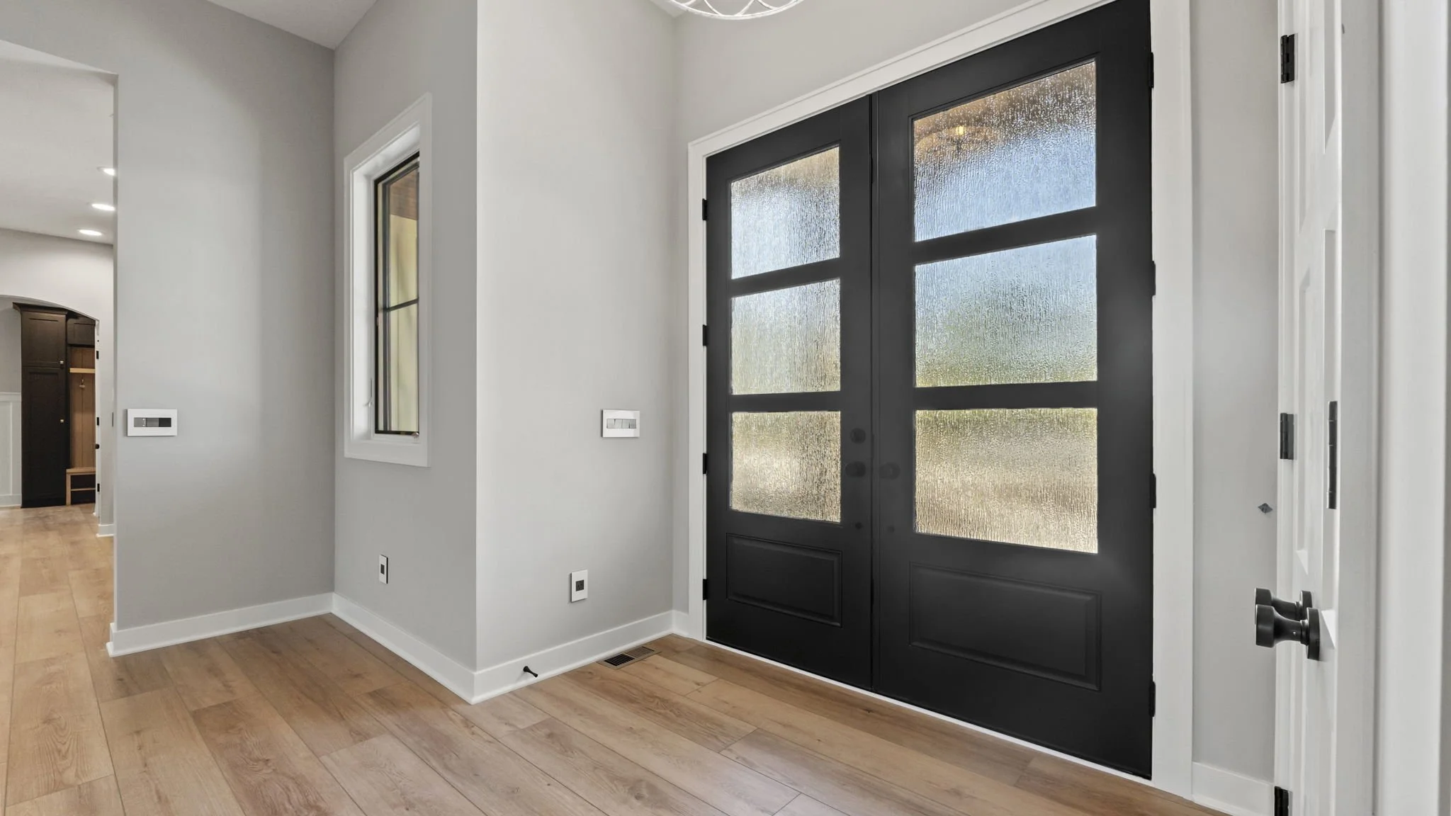 Entryway with black front door with frosted glass panels, white walls, medium-tone wood flooring, and a small vertical window on the wall.