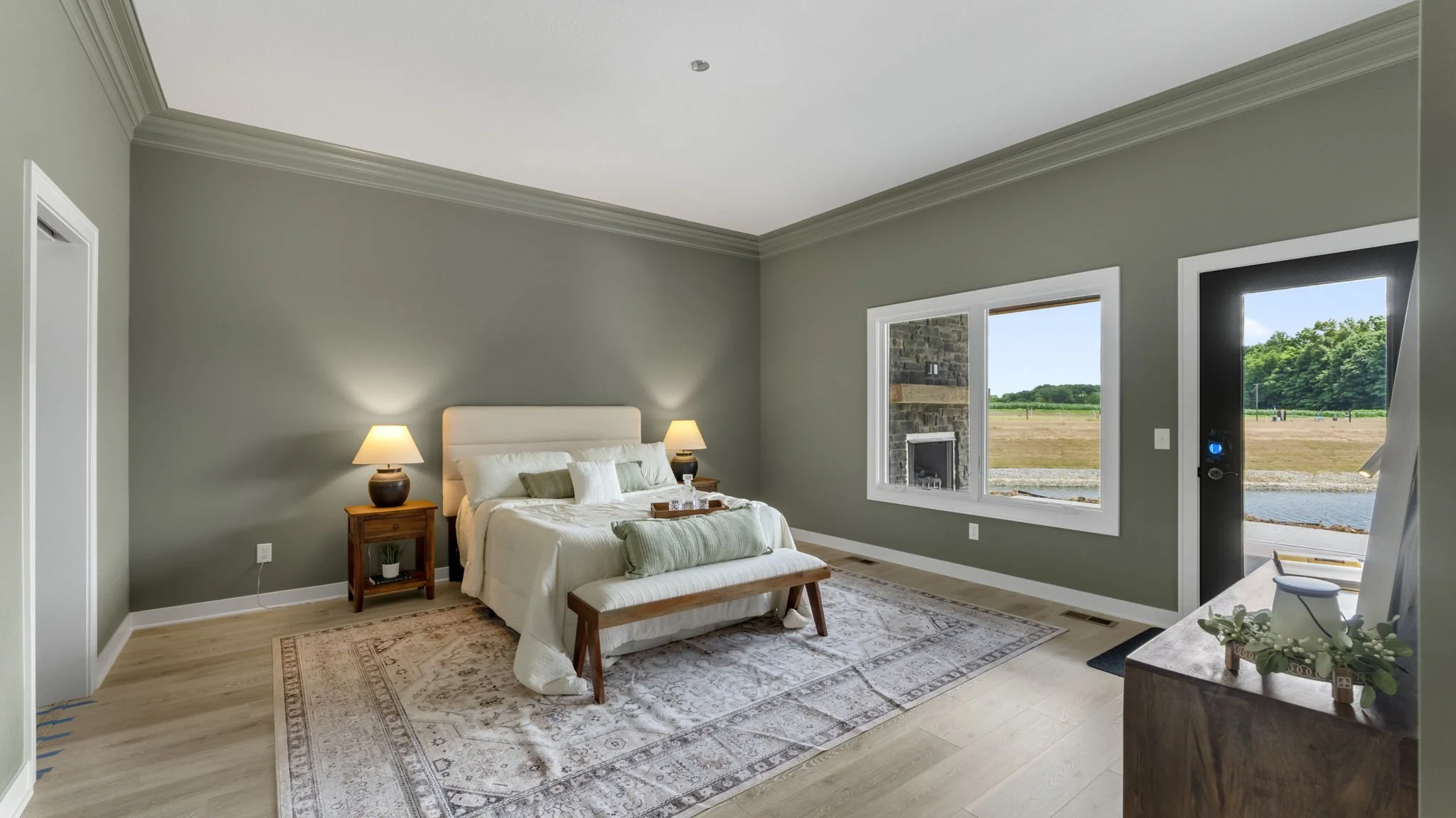 Modern bedroom with light green walls, a large bed with white bedding and pillows, two bedside tables with lamps, a bench at the foot of the bed, large windows showing outdoor landscape, and a view of a fireplace outside the window.