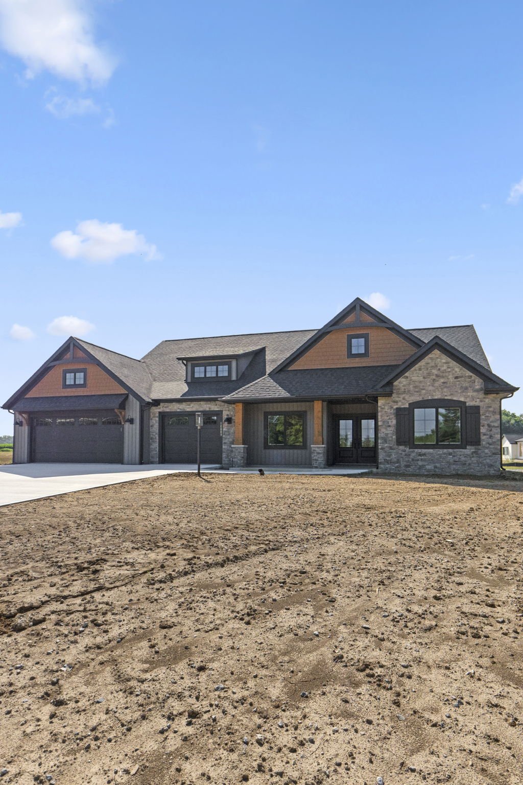 Newly built house with a mix of stone and wood siding, large windows, a big garage, and a landscaped front yard under a blue sky.