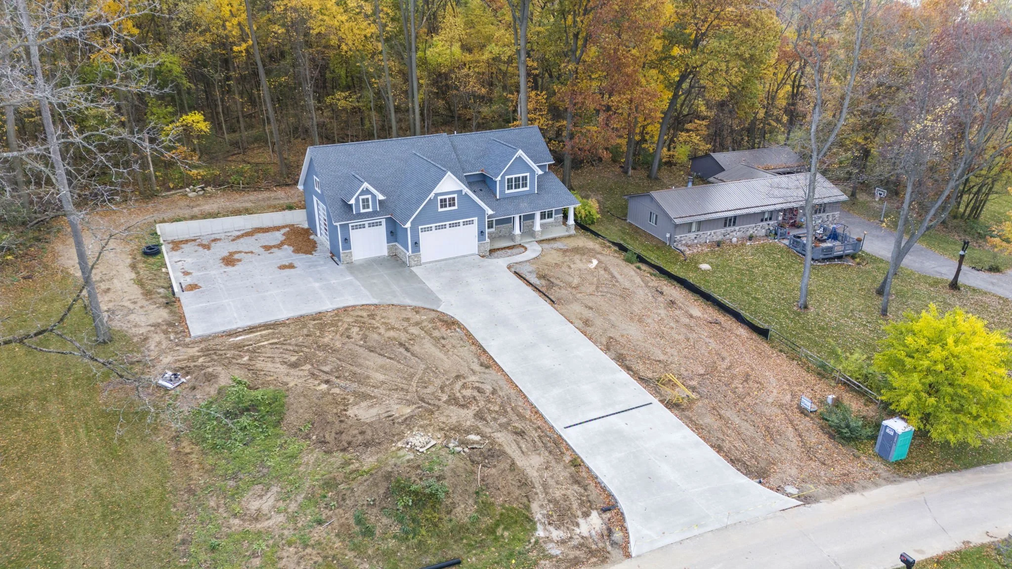 A house under construction with a large concrete driveway, surrounded by trees with fall foliage, and a portable toilet nearby.