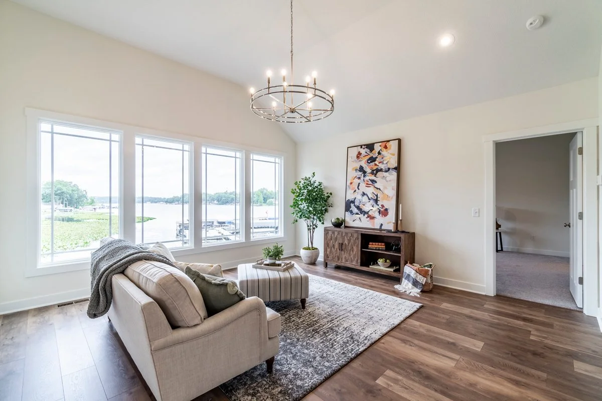 Living room with large windows, beige sofa, ottoman, abstract artwork, wooden media console, potted plant, chandelier, and carpet on hardwood floor.