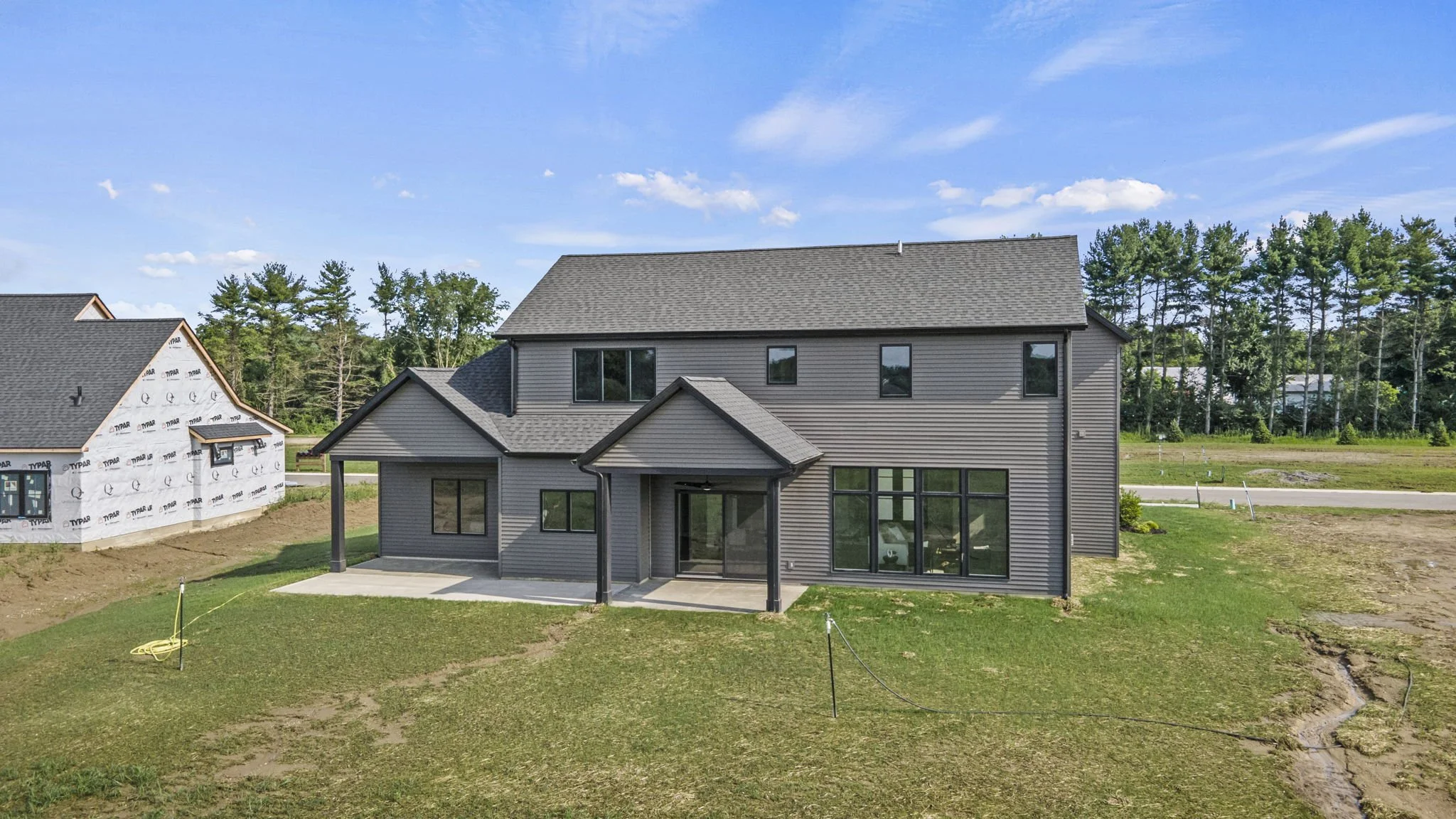 Newly constructed gray two-story house with black window frames, a covered porch, and a backyard with grass and a concrete patio. There is a neighboring house under construction on the left and a tree line in the background. The sky is clear with a f