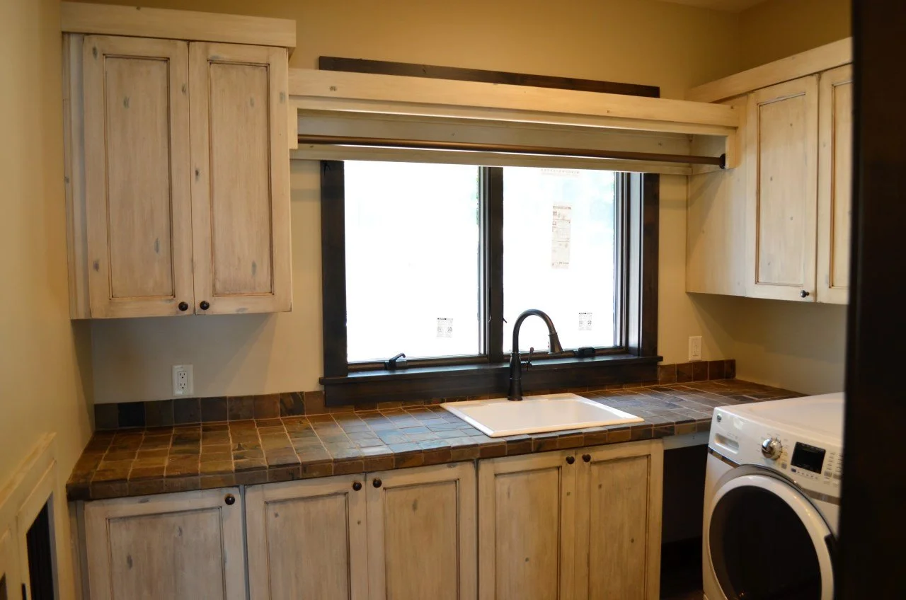 A laundry room with light wood cabinets, a black faucet, a white sink, a window with black trim, a brown tiled countertop, and a washing machine.