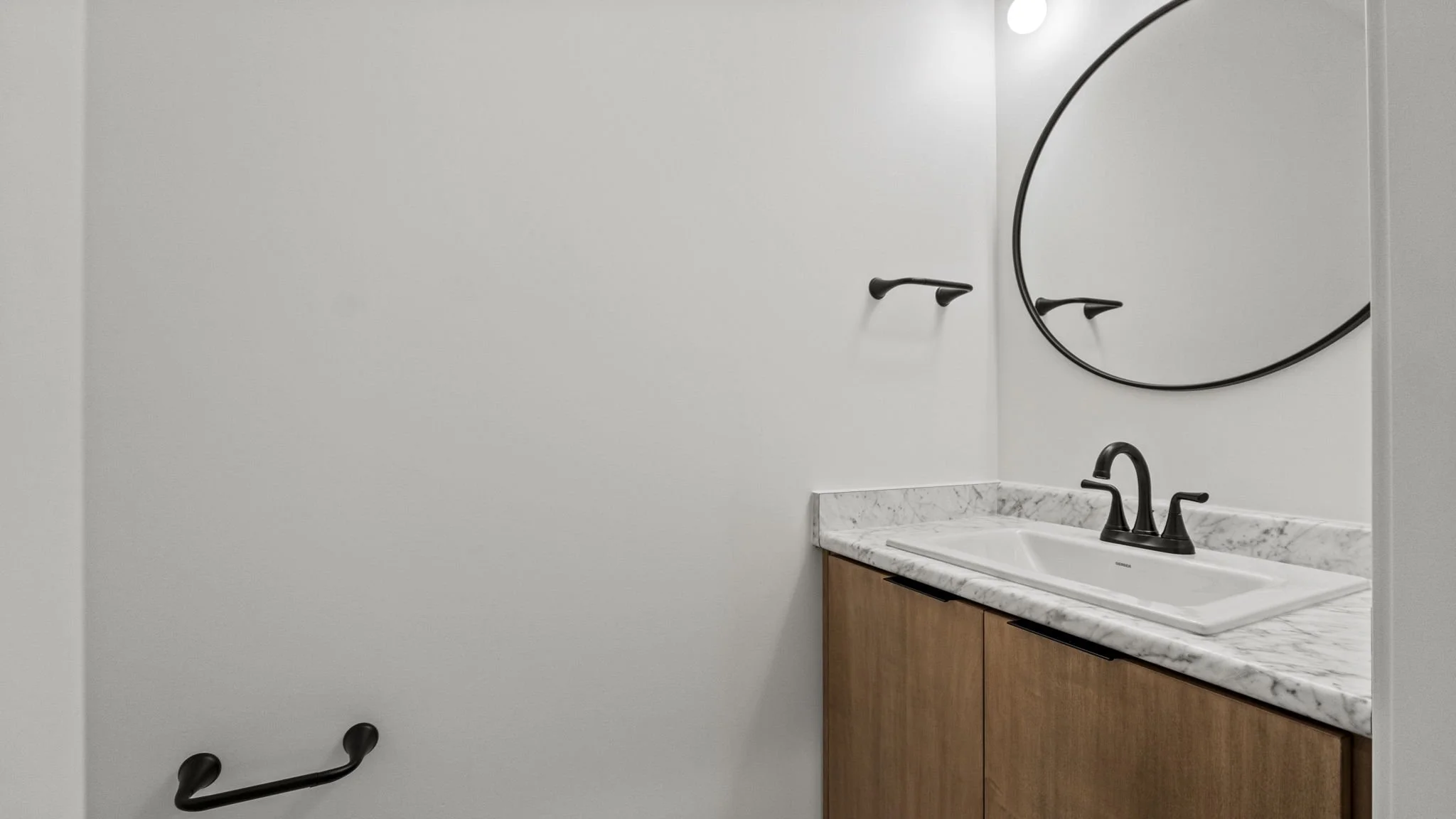 Bathroom with a marble countertop, rectangular sink, black faucets, round mirror, and black wall-mounted towel hooks on a white wall.