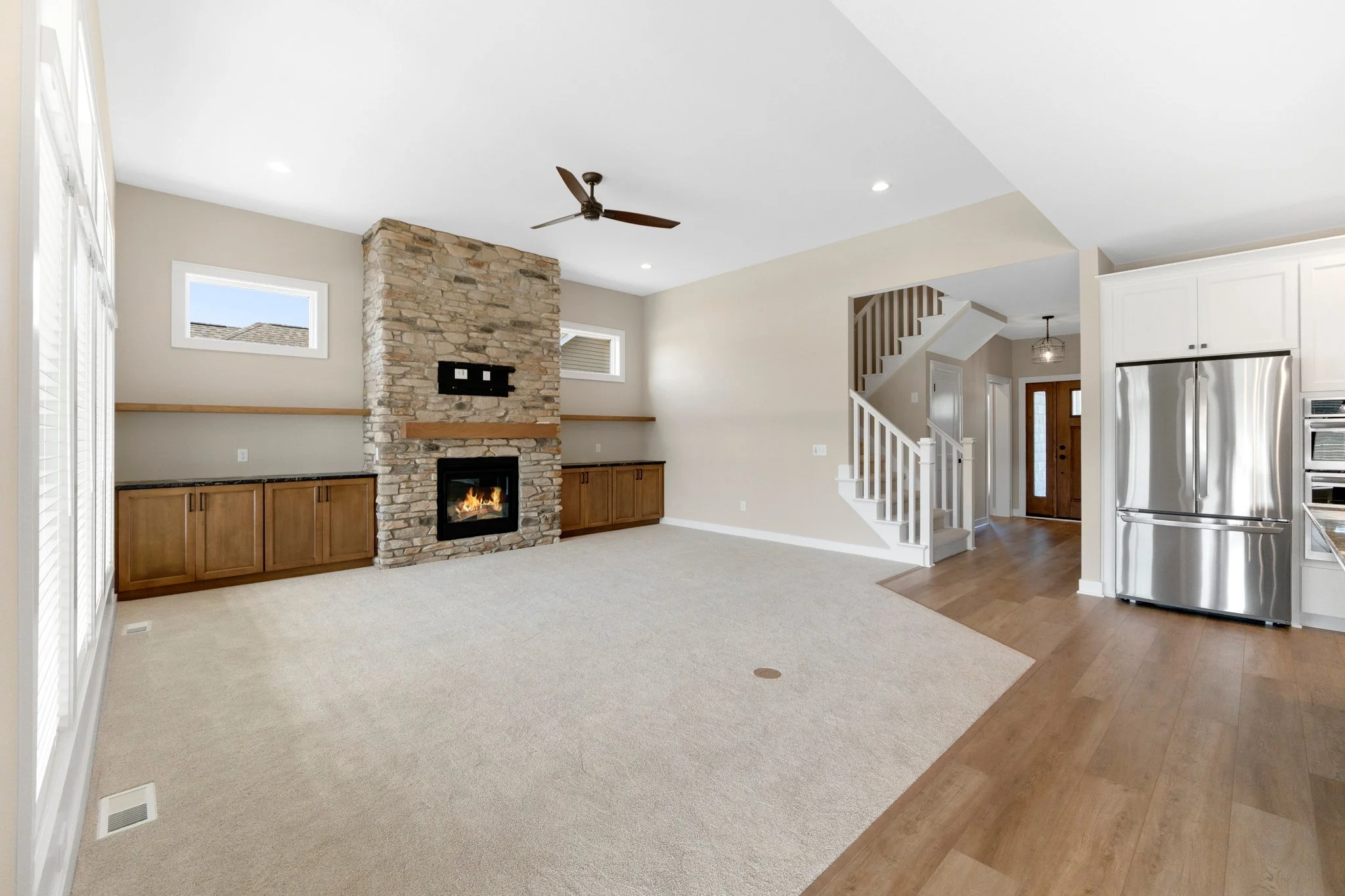 Living room with stone fireplace, beige carpet, built-in wooden cabinets, white staircase, adjacent kitchen with stainless steel refrigerator, and wooden flooring near the entrance.