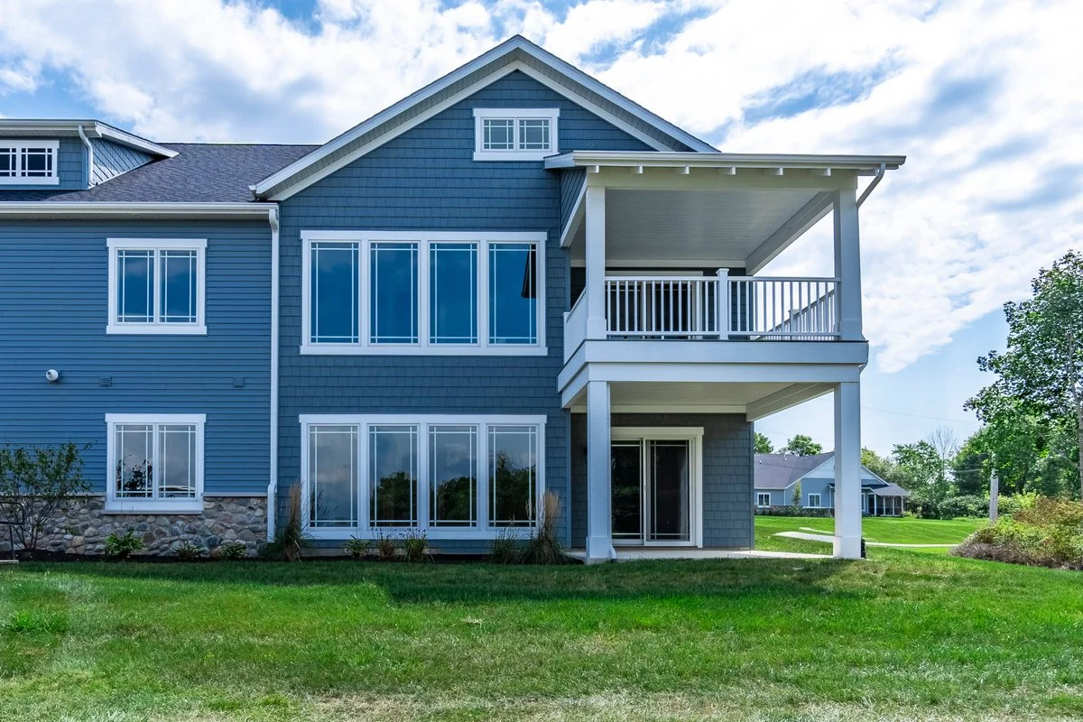 A modern, multi-story house with blue siding and white trim, featuring large windows and a spacious balcony with white railings, situated on a well-maintained grassy lawn under a partly cloudy sky.
