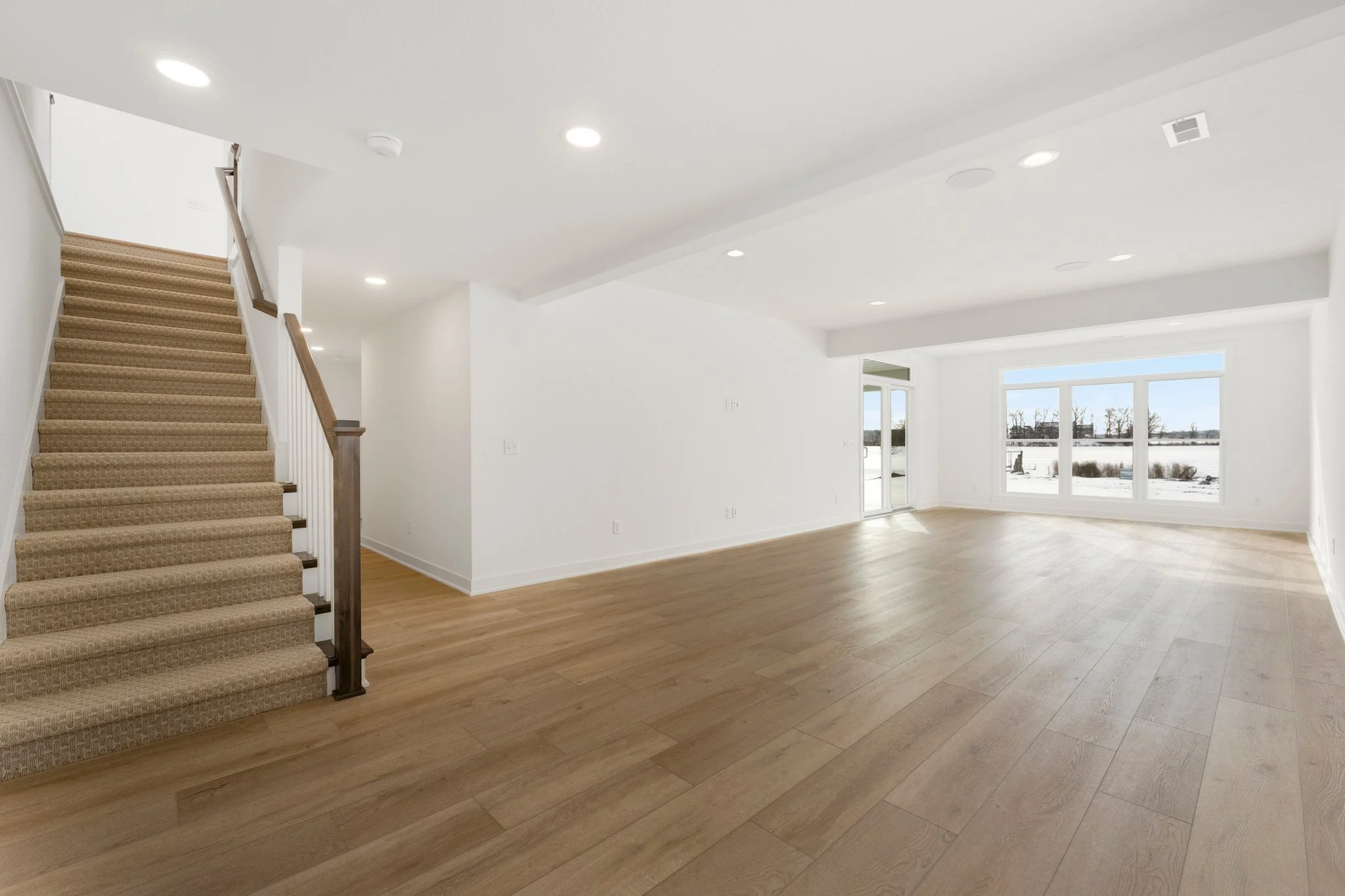 Empty living room with hardwood floors, large windows showing snowy landscape, and a staircase with beige carpet leading upstairs.